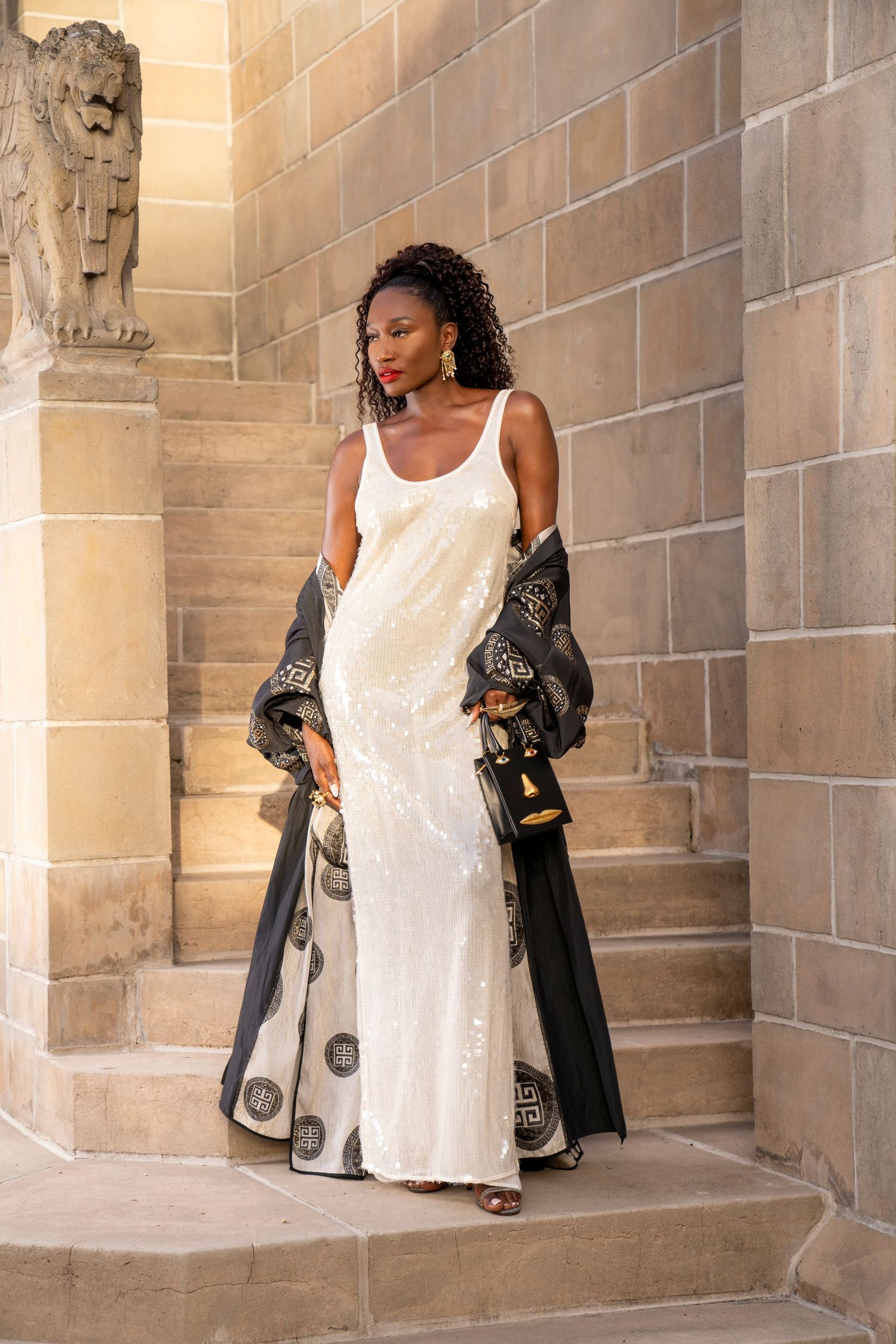 Lifestyle modeling photo of black woman standing with statue and staircase behind her. Woman in Upscale white dress and cover.