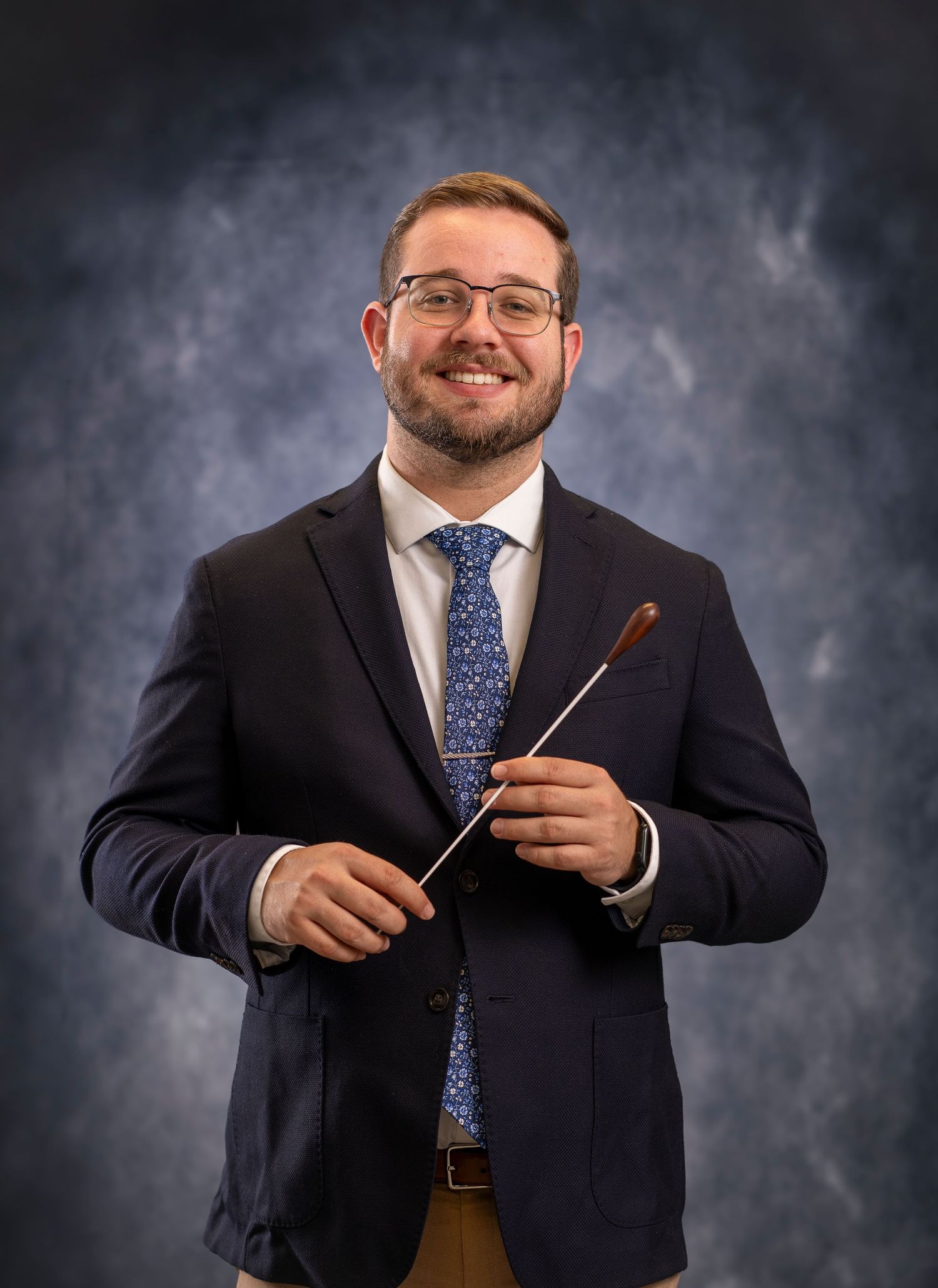 School headshot portrait of band director holding baton in grey faux background.