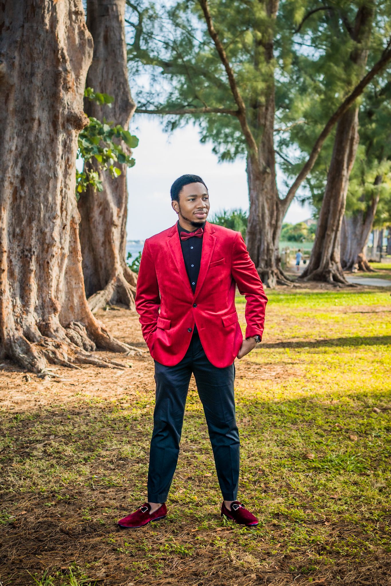 black man in red suit and black pants posing in forest by Large trees with greenery and trees out of focus in background