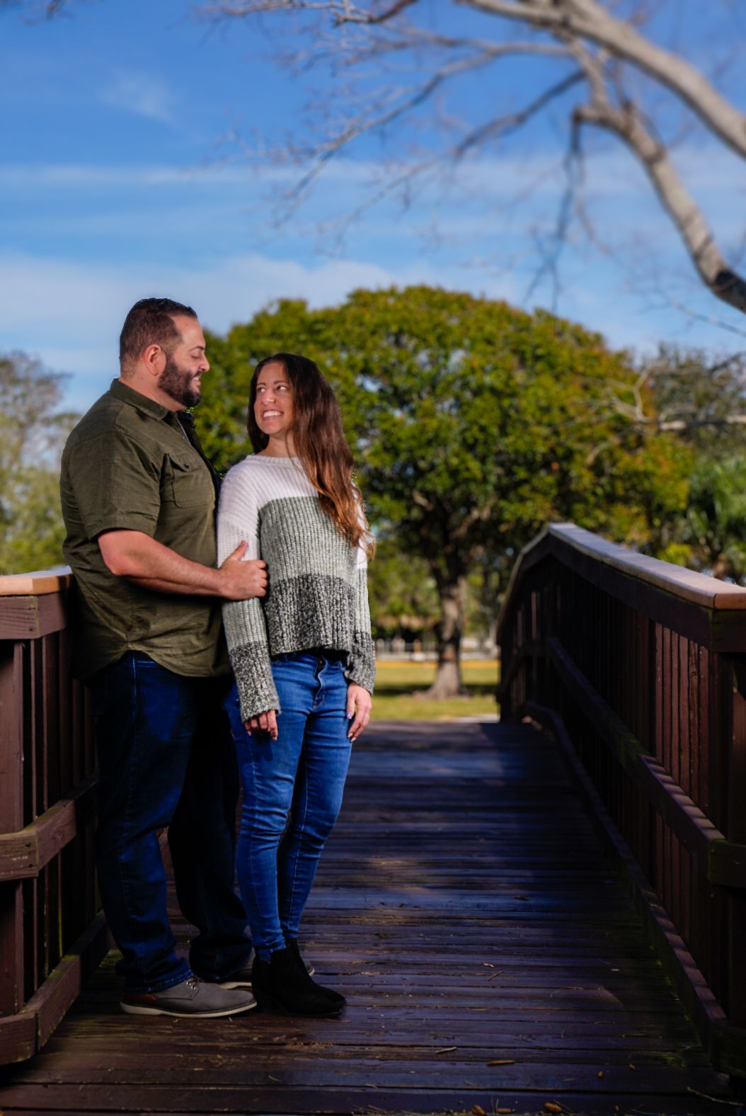 Man and woman standing up against bridge with green trees in background. Man holding onto woman. Woman looks back at man. Both gazing.