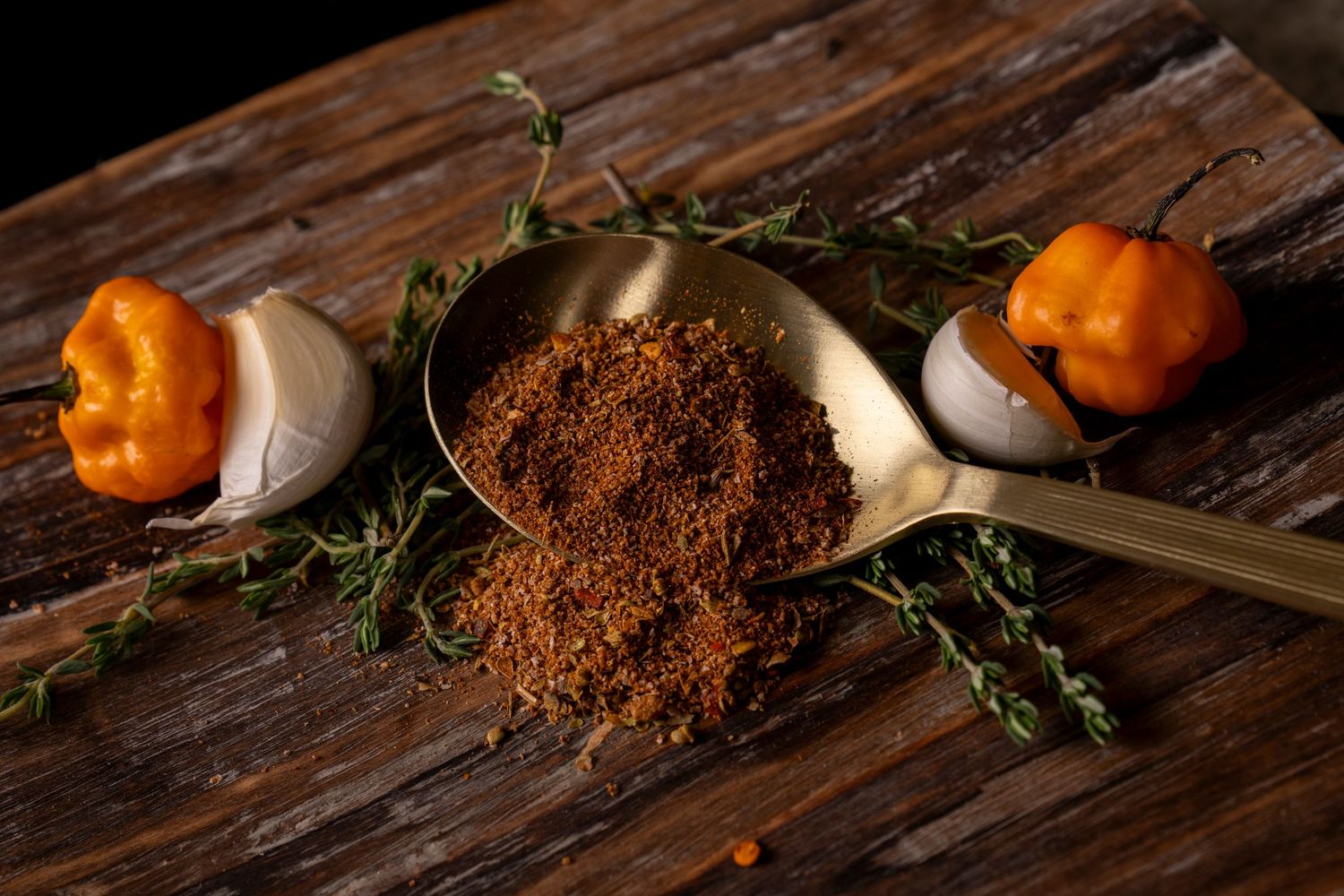 Food photography close up of spices on spoon sitting on fresh herbs and peppers on brown textured cutting board.