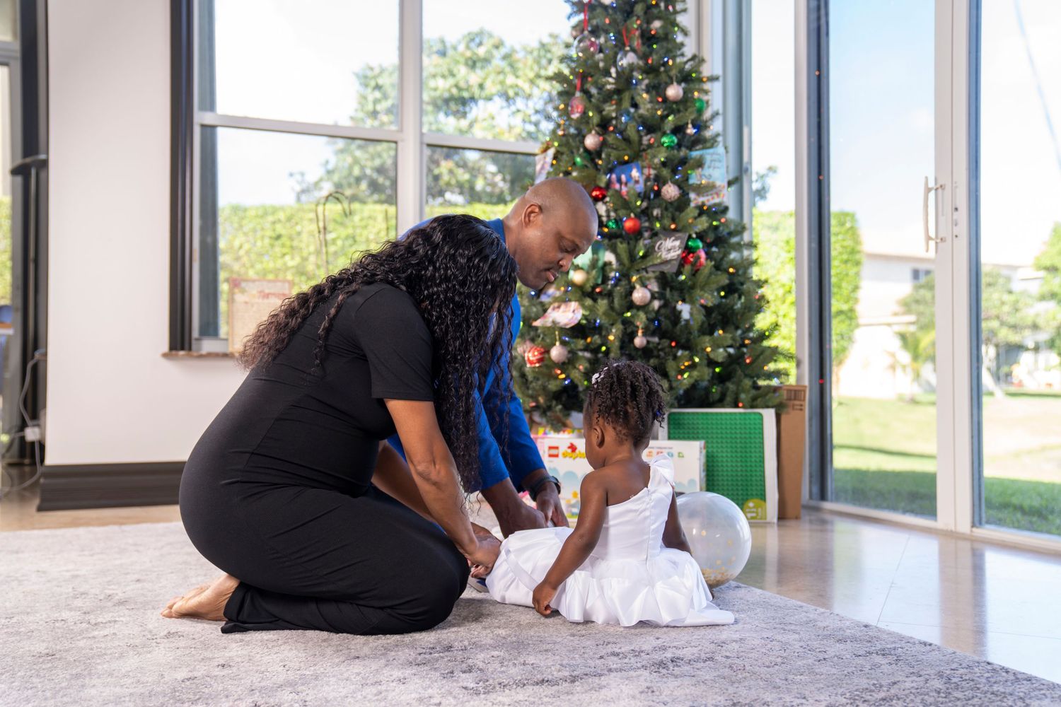 Mother and father in house fixing 2 year old's white dress while christmas tree in background.