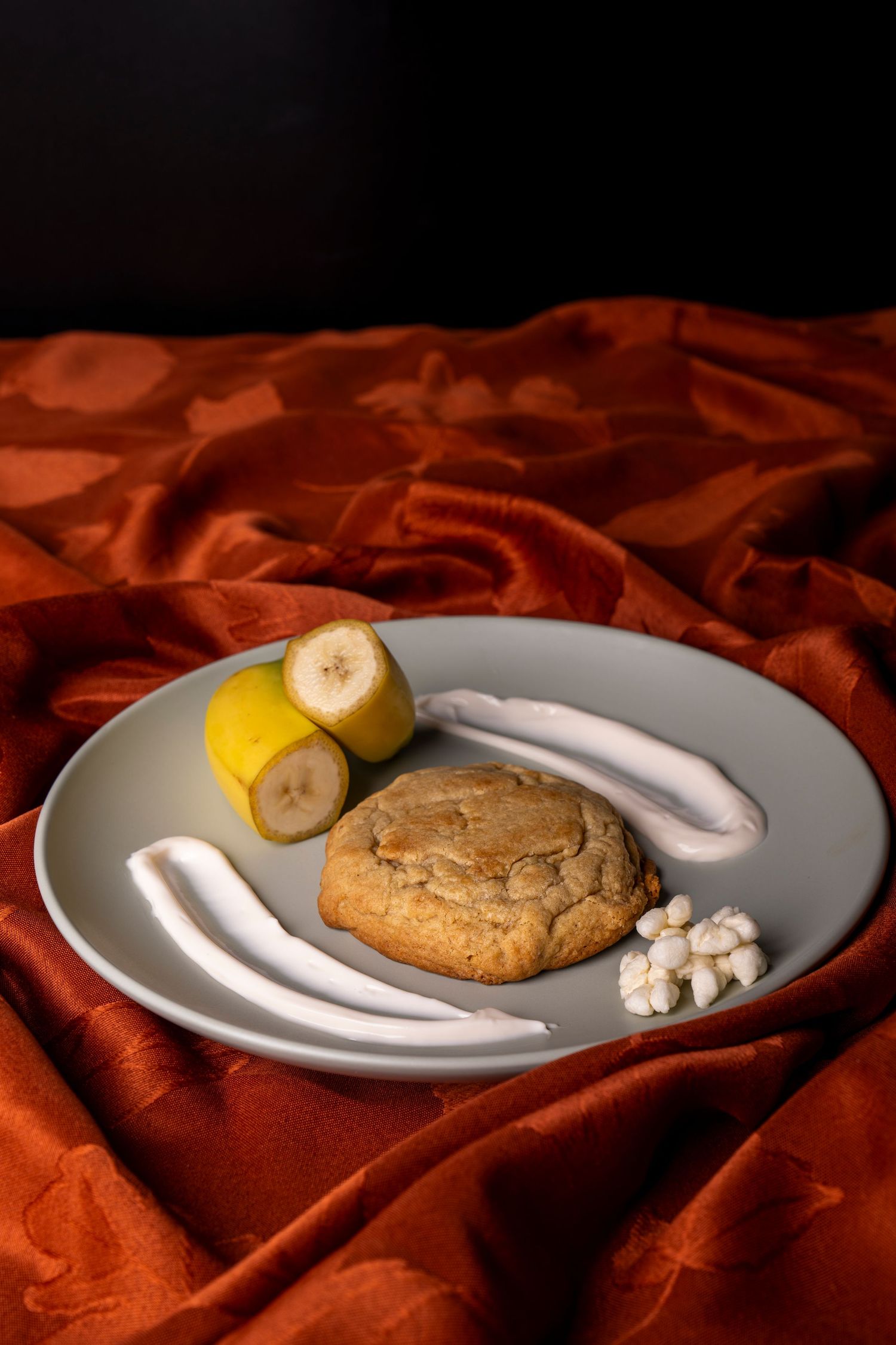 Food photography. Cookies displayed on round plate with sugar, bananas, and marshmallow. Sitting on crinkled red fabric.