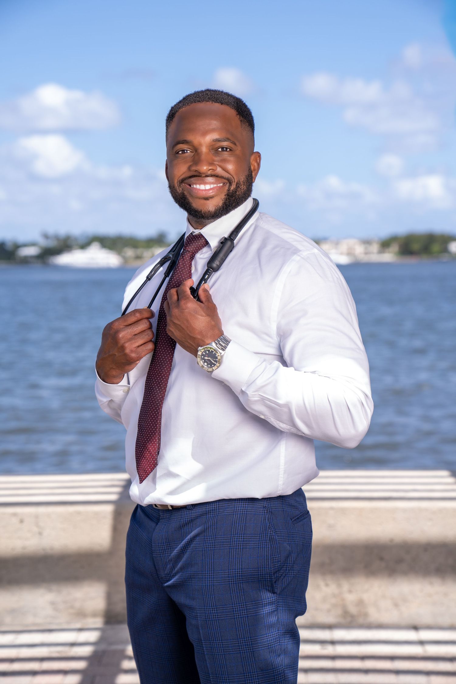 Doctor's headshot branding photo in West Palm Beach Florida. Holding stethoscope around neck, smiling, and city and blue skies in background