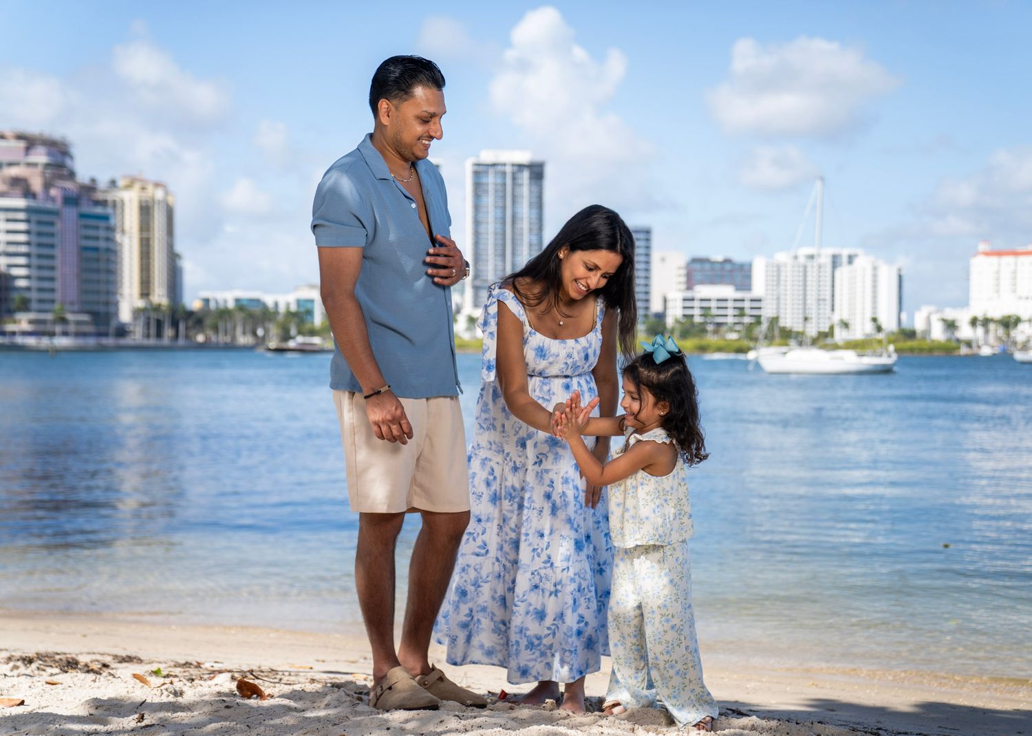 Pregnant mother, father and daughter posing for family photos on beach with West Palm Beach City in the background. Palm Beach Florida