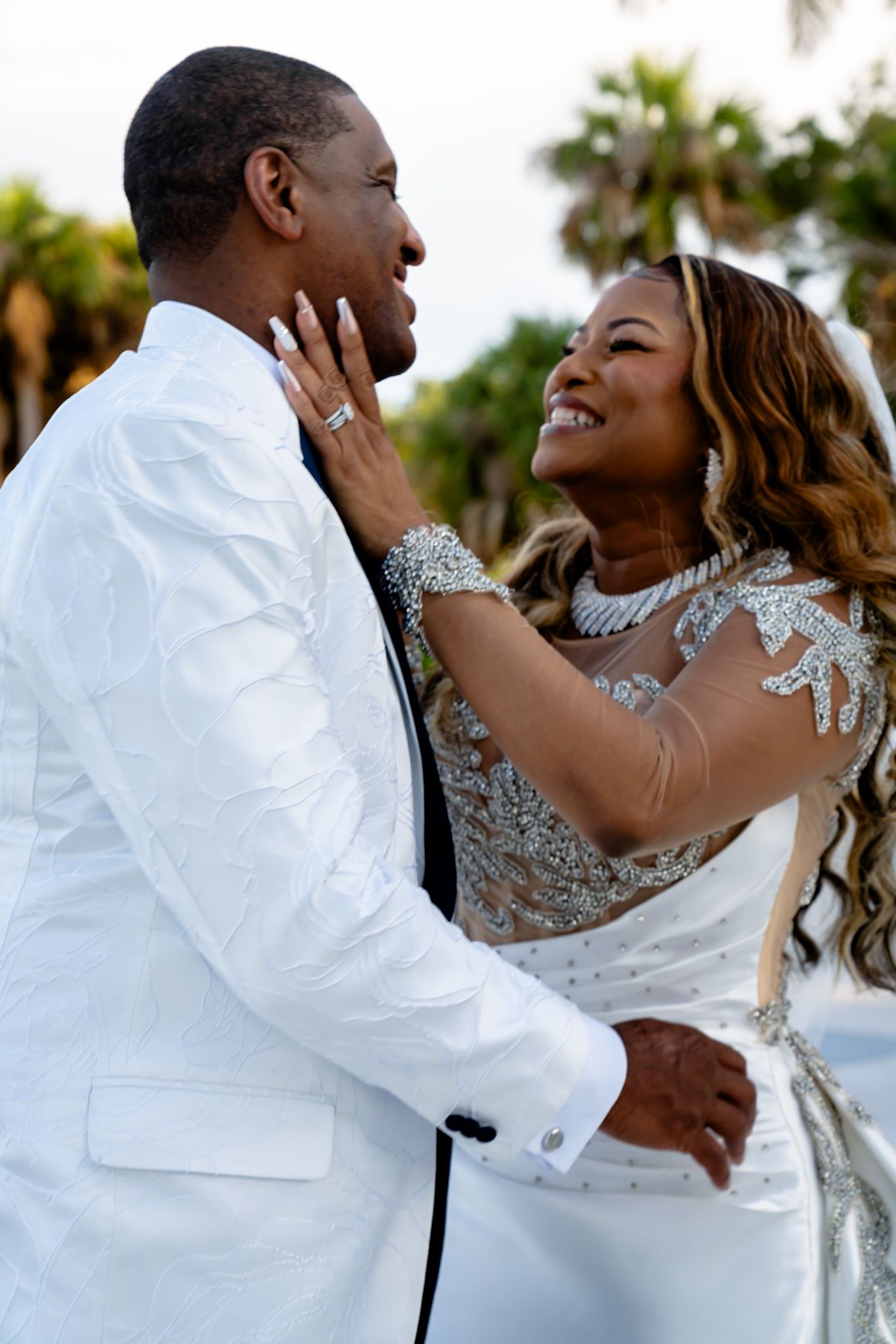bride caresses grooms faces as they smile and gaze into each other's eyes. close up shot with greenery in blur in background.