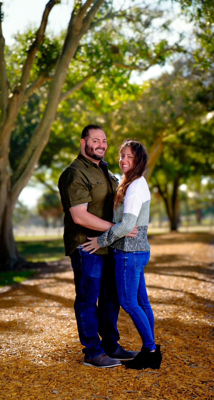 man and woman hold each other by waste in forest while smiling. blurred greenery in backgornd. Standing on woodchipped path.