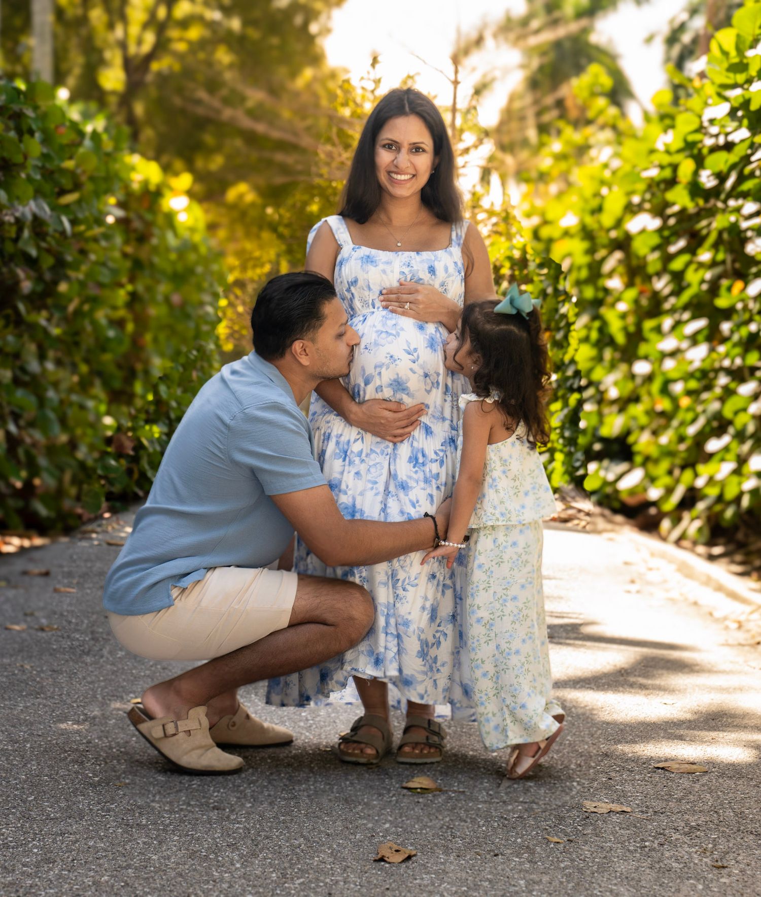 Maternity photoshoot with father and daughter kissing mother's belly while posing in greenery background. Palm Beach Florida