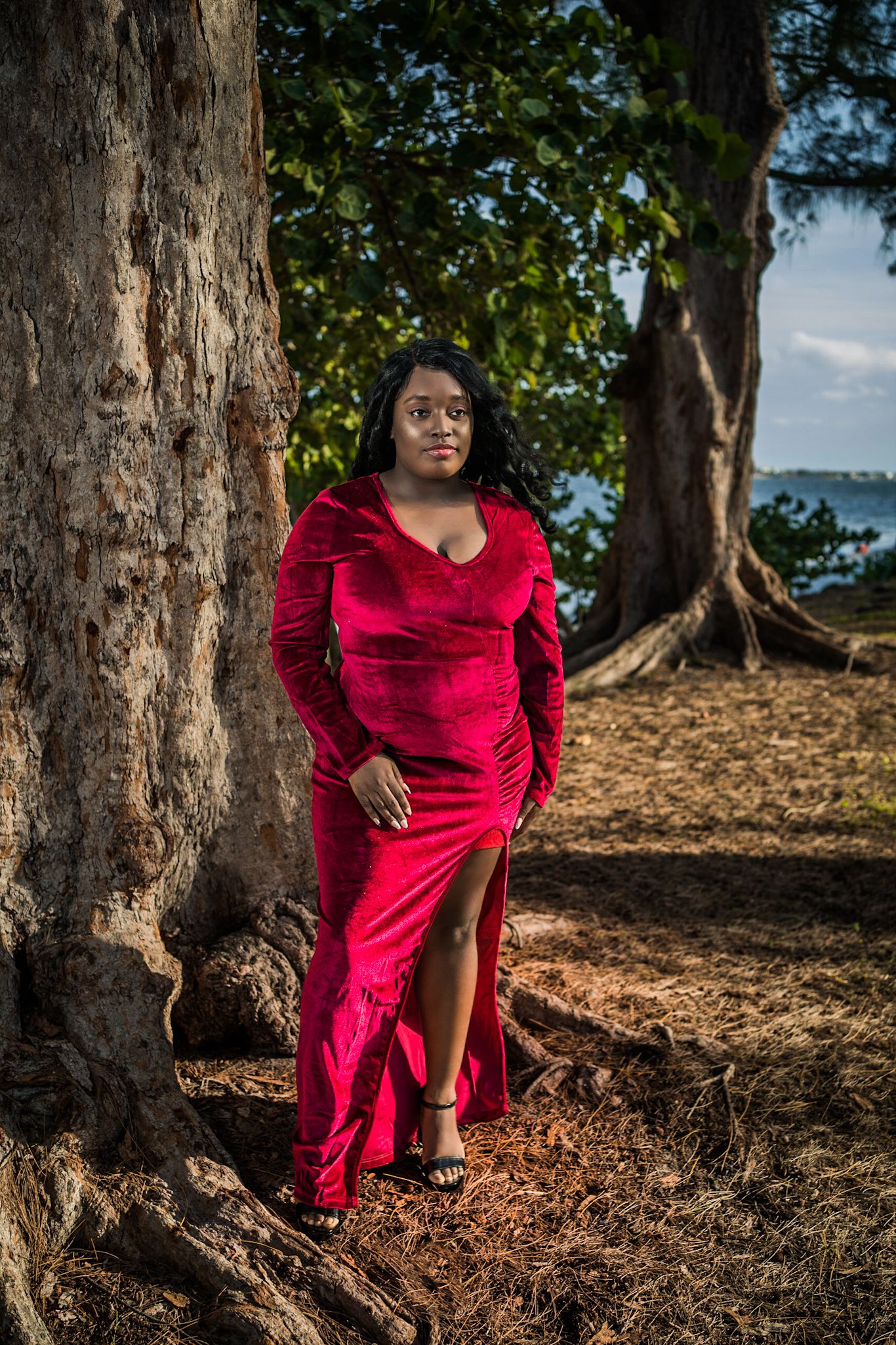 black Woman in red posing in forest by Large trees with greenery and trees out of focus in background