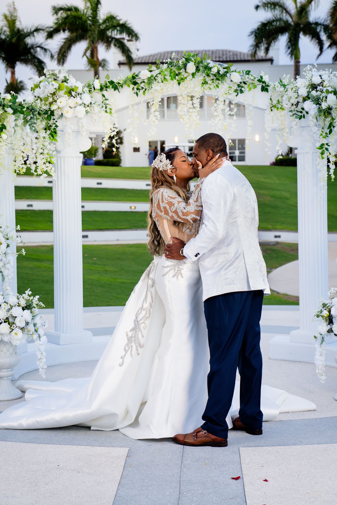 Bride and groom kiss under floral and greenery-lined archway with mansion in bakcround.
