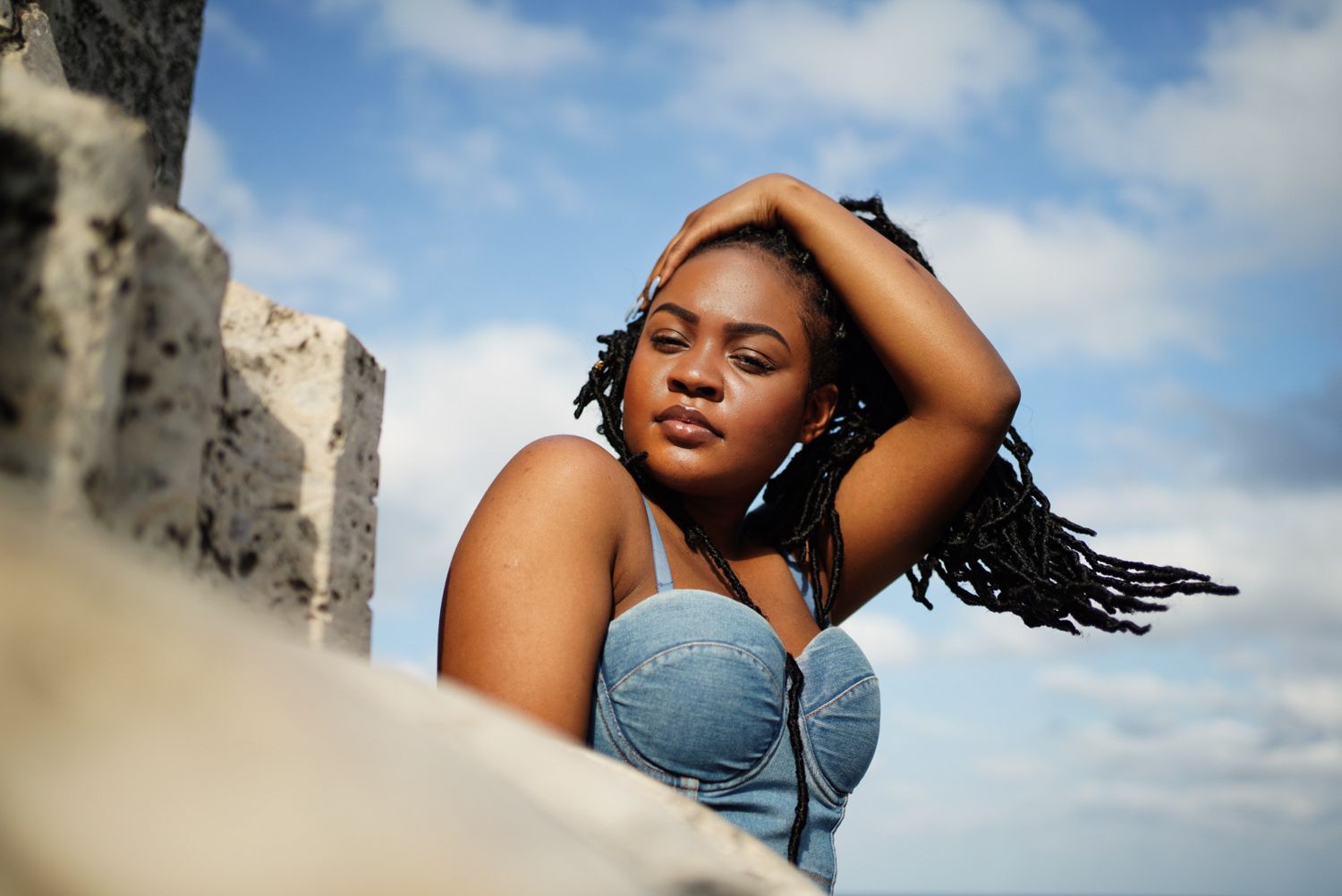 Black woman with braided hair blowing in wind. Artistically posing with hand on head, coral architecture i foreground, and blurred skies.