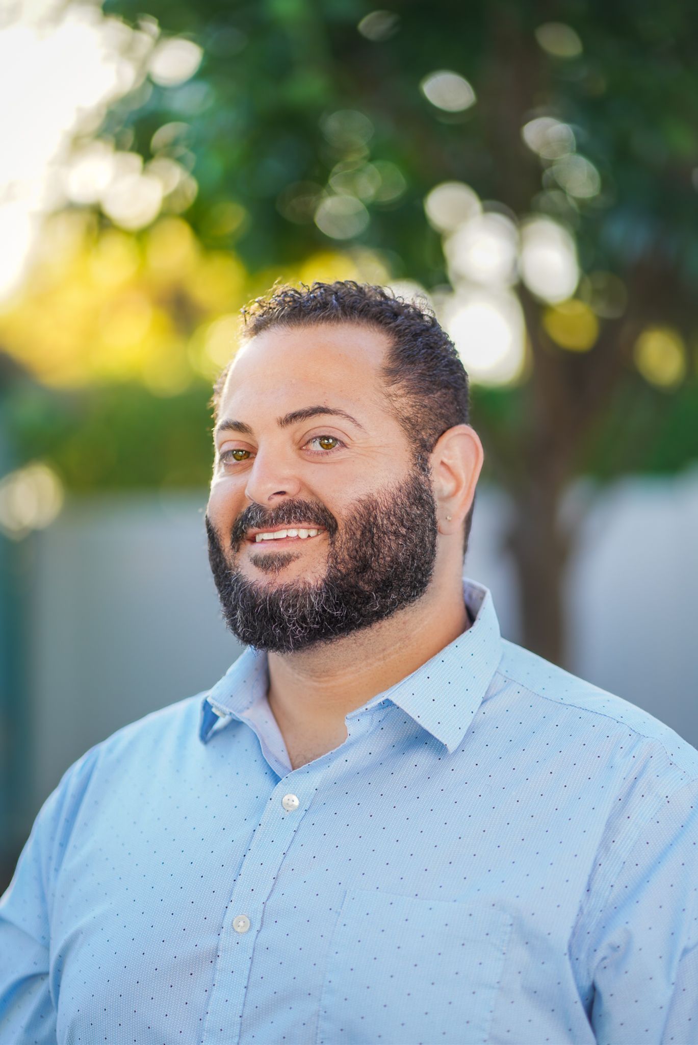 Headshot portrait of man with beard smiling in blue shirt with trees and white wall background.