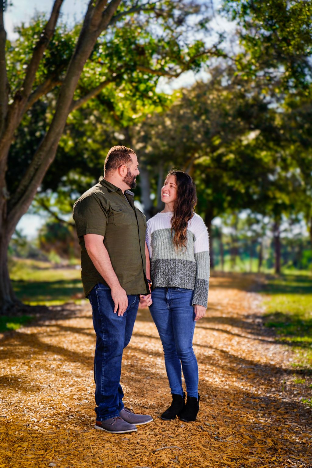 Couple on woodchipped path and greenery in background out of focus. Couple holding hands and smiling while looking at each other.