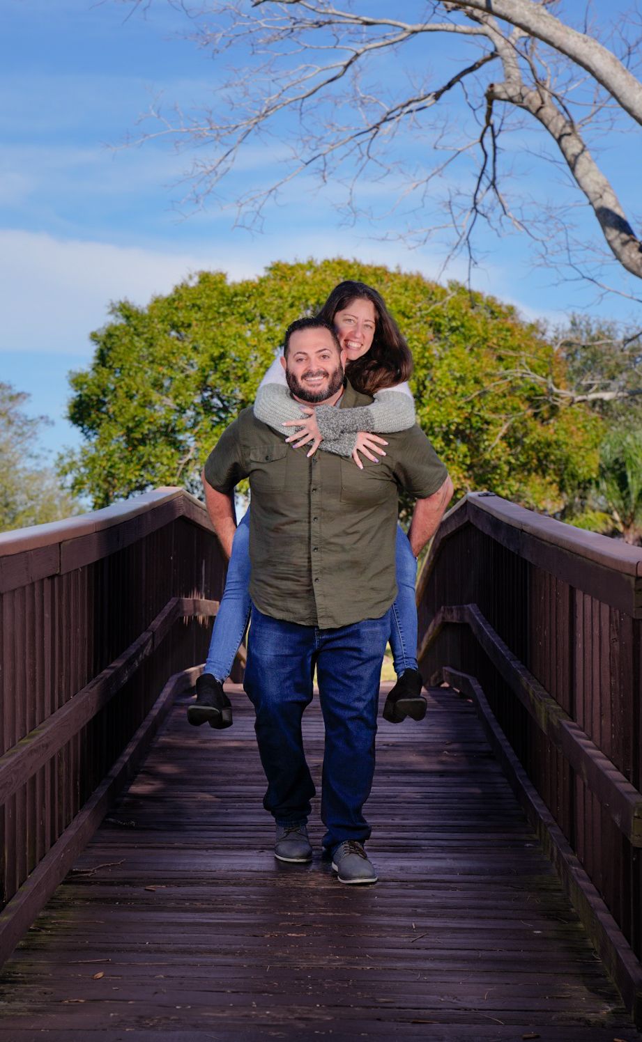 man gives piggyback ride to woman on brown bridge with greenery and blue skies blurred in background.