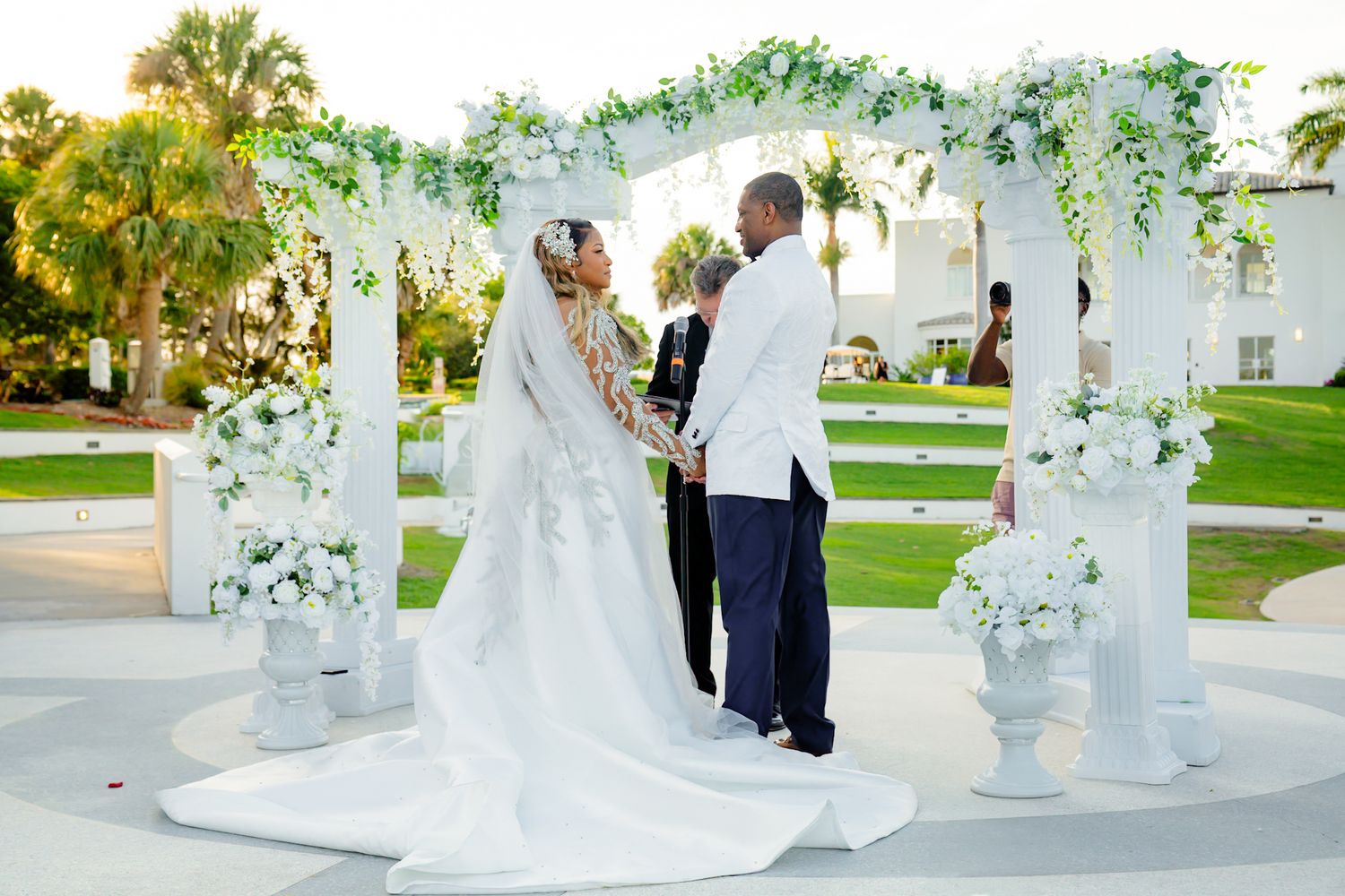 bride and groom hold hands. officiant marries them under white columned archway lined with flowers and greenery. mansion and trees behind