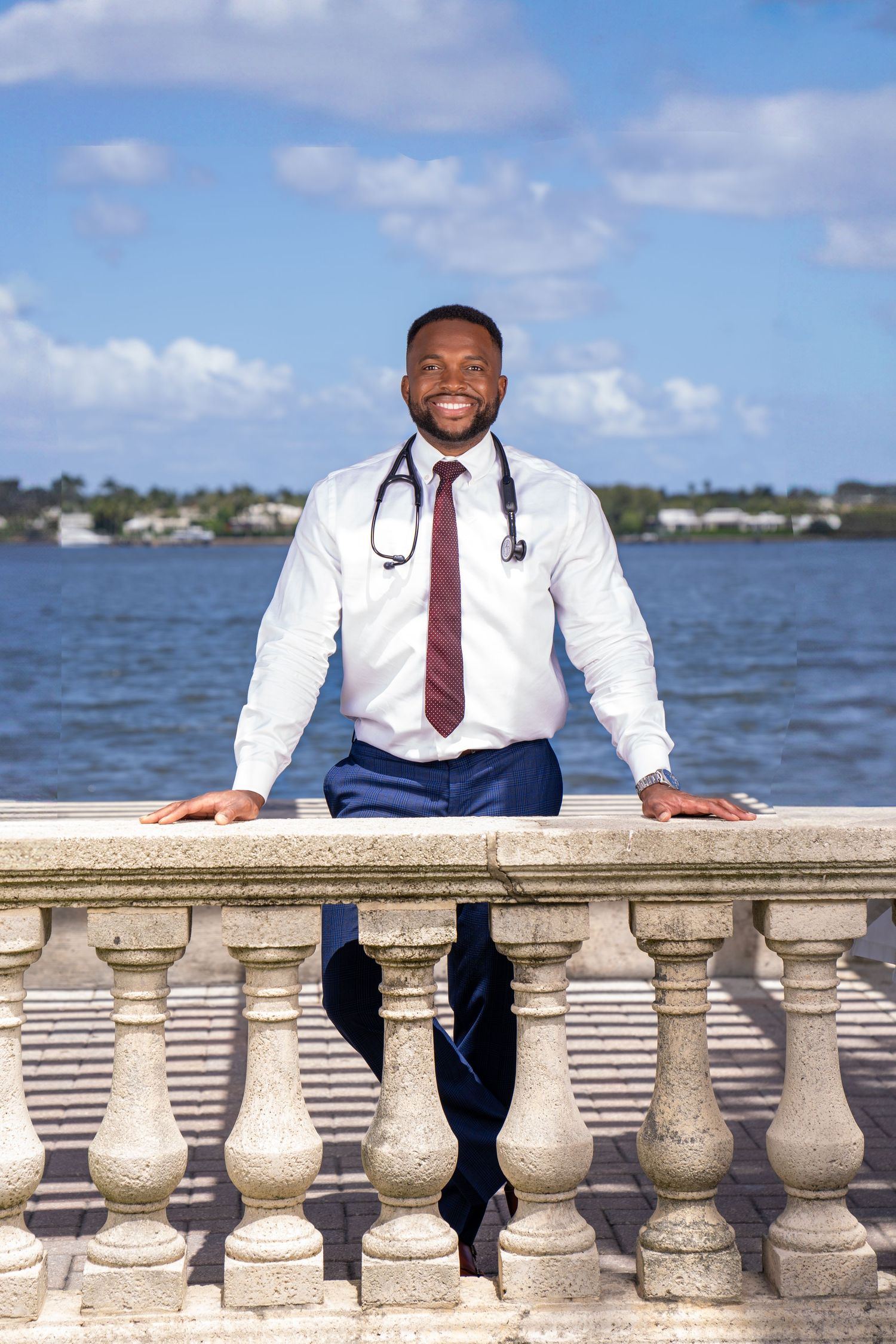 doctor poses with hands on choral barrier with stethoscope on neck and blue skies, clouds,  and cityscape in background.
