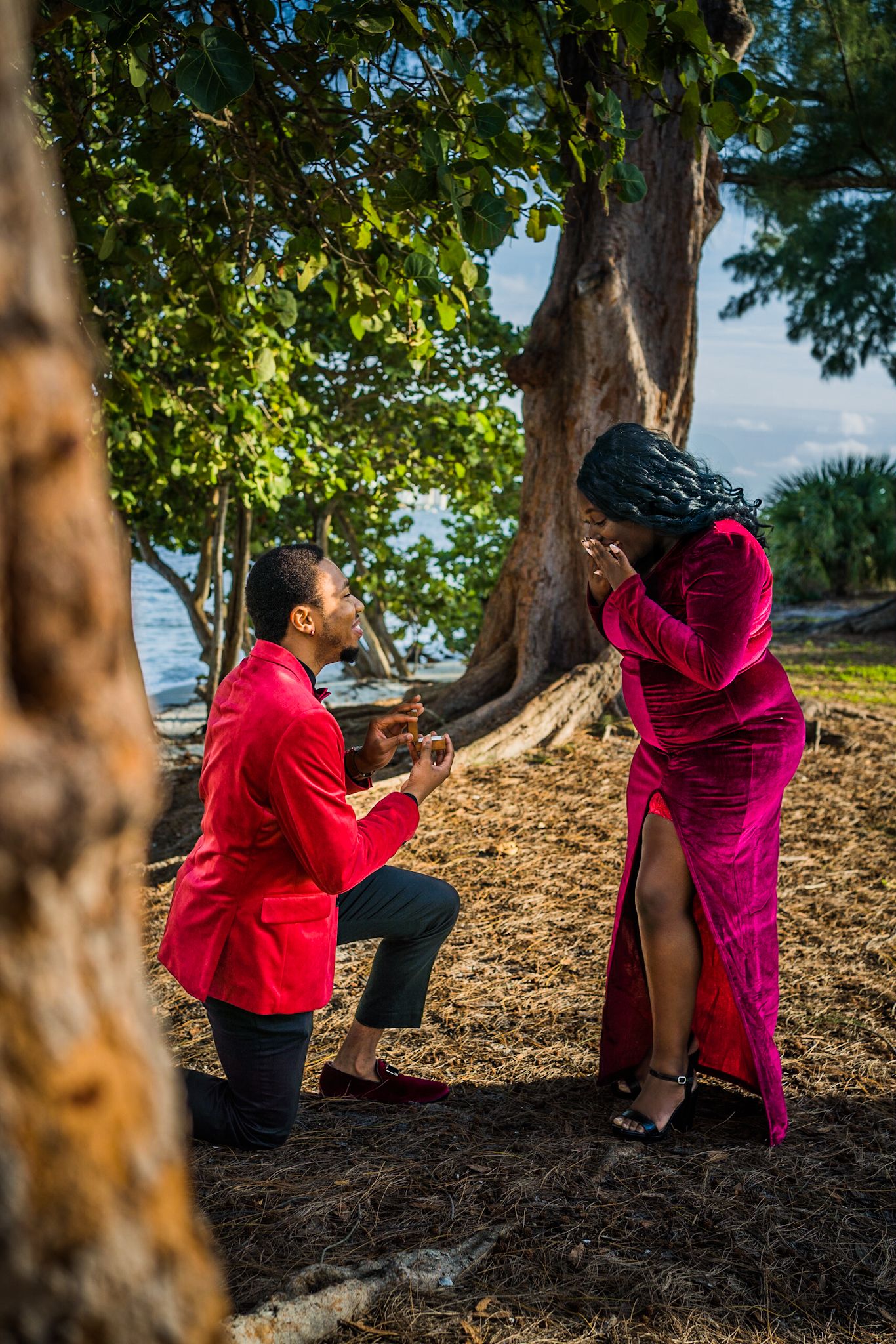 Man in red suit proposing to woman in red dress in forest with water in background. Woman hands over her face surprised.
