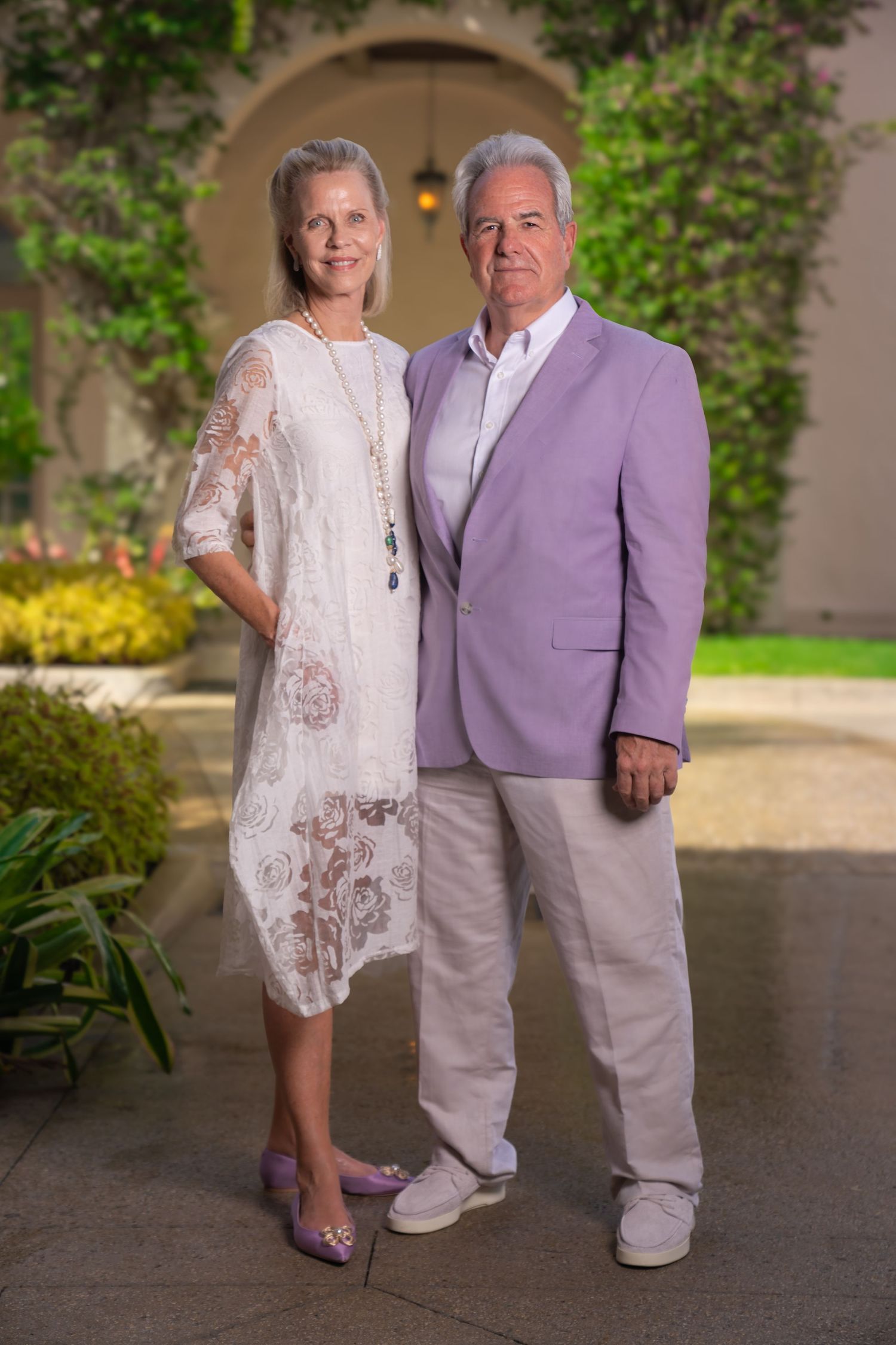 Older couple portrait on palm beach island with archway, greenery, and blurred background. man in lavender suit and woman in white dress.