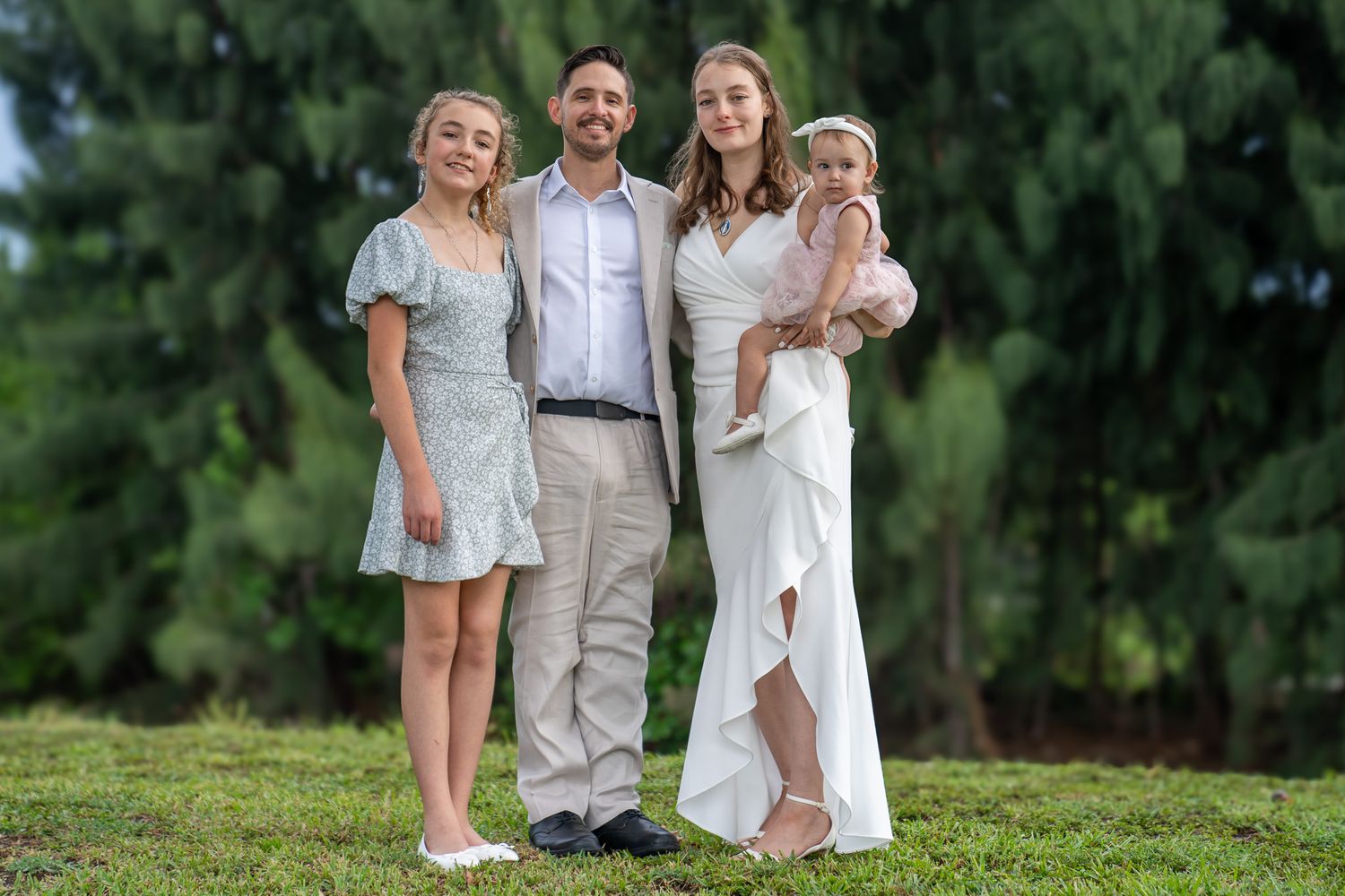 Family posing in Forest setting just after marriage. Wife in wedding dress, daughter in green dress, husband in tan suit, and baby in pink.