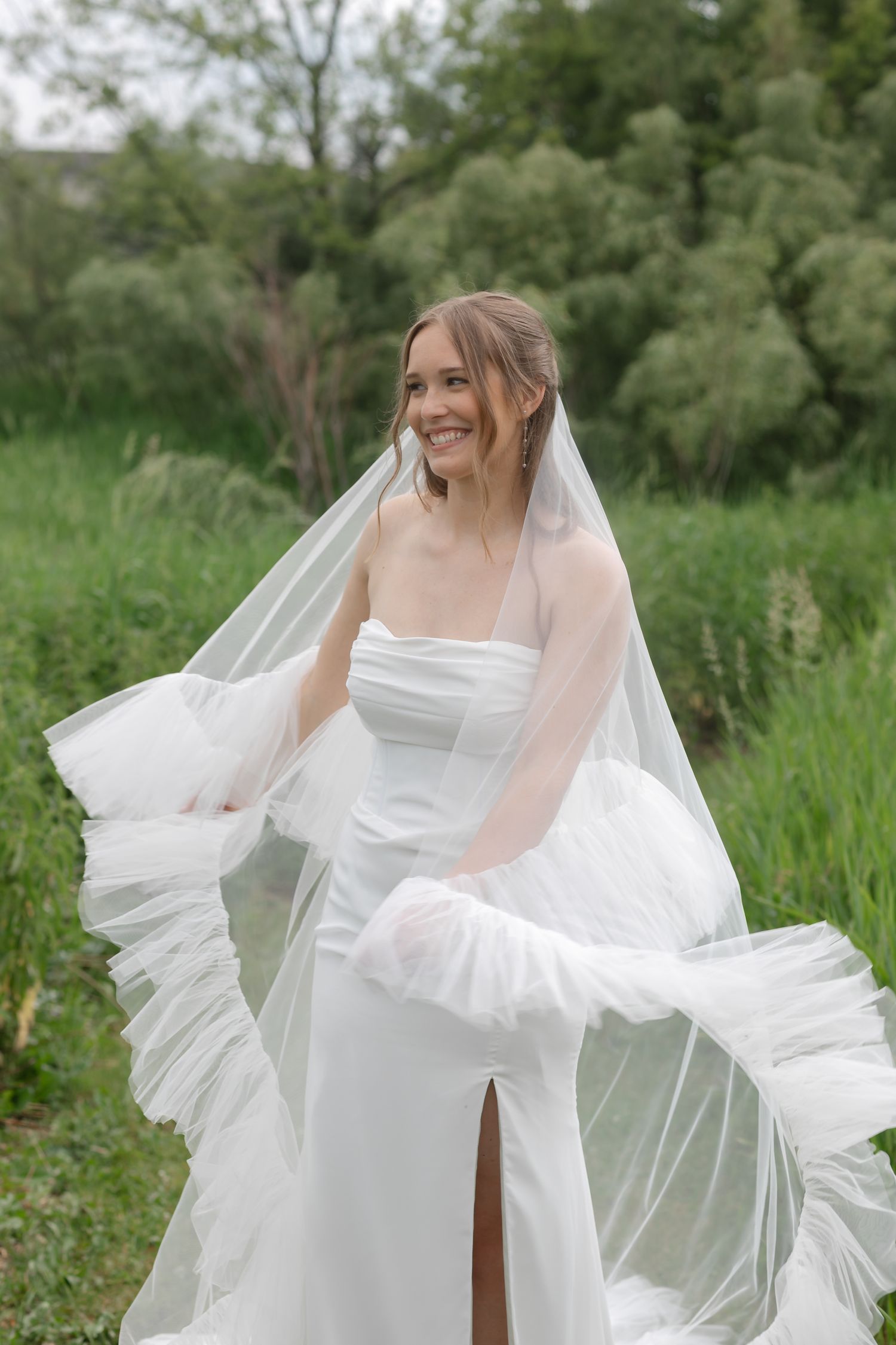 Smiling bride in white strapless gown with veil and gloves posing outdoors in lush green garden setting.