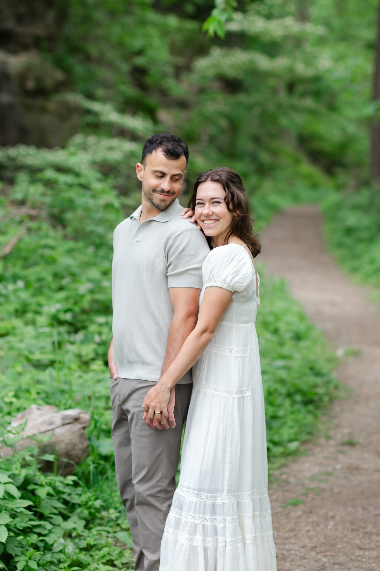 A smiling couple holds hands on a lush green forest path, woman in white dress, man in gray polo shirt.