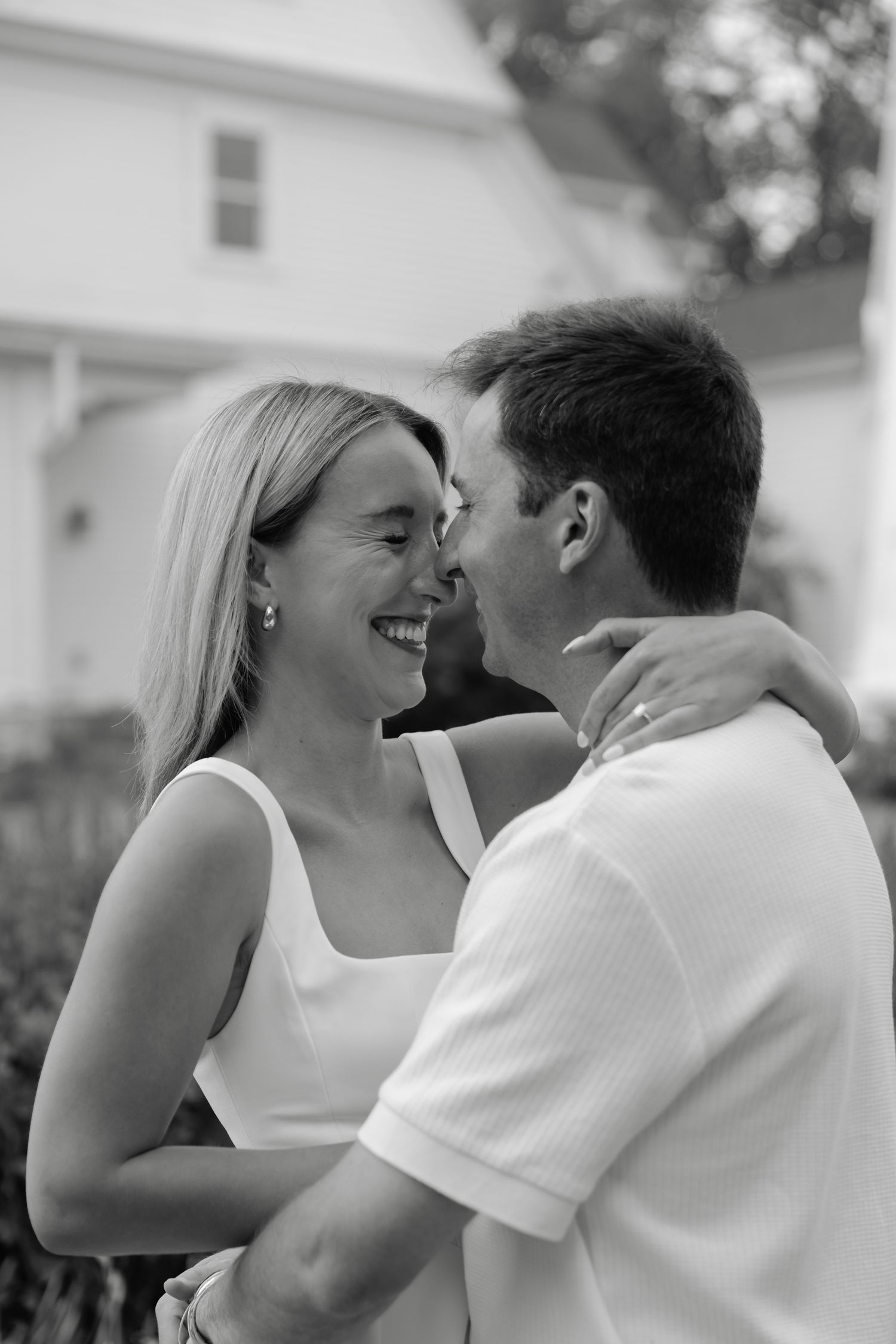 Black and white photo of a smiling couple embracing outdoors near a white house.