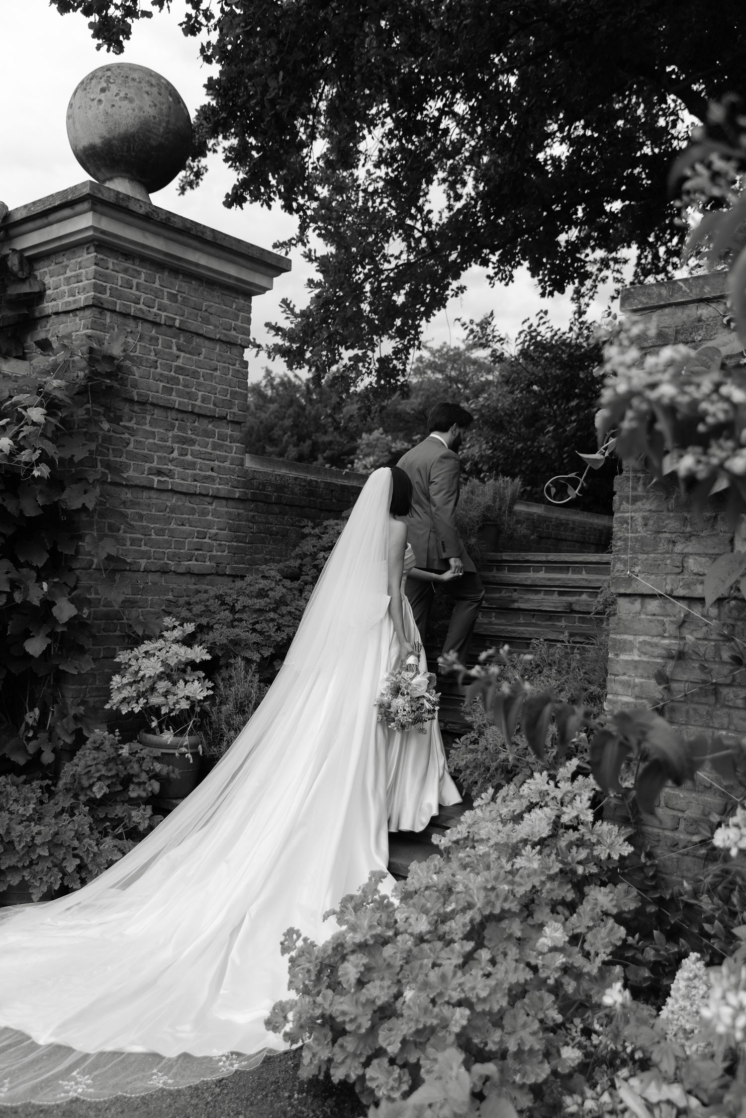 Black and white photo of a bride and groom entering a garden gate, her long veil trailing behind amid lush flowers.