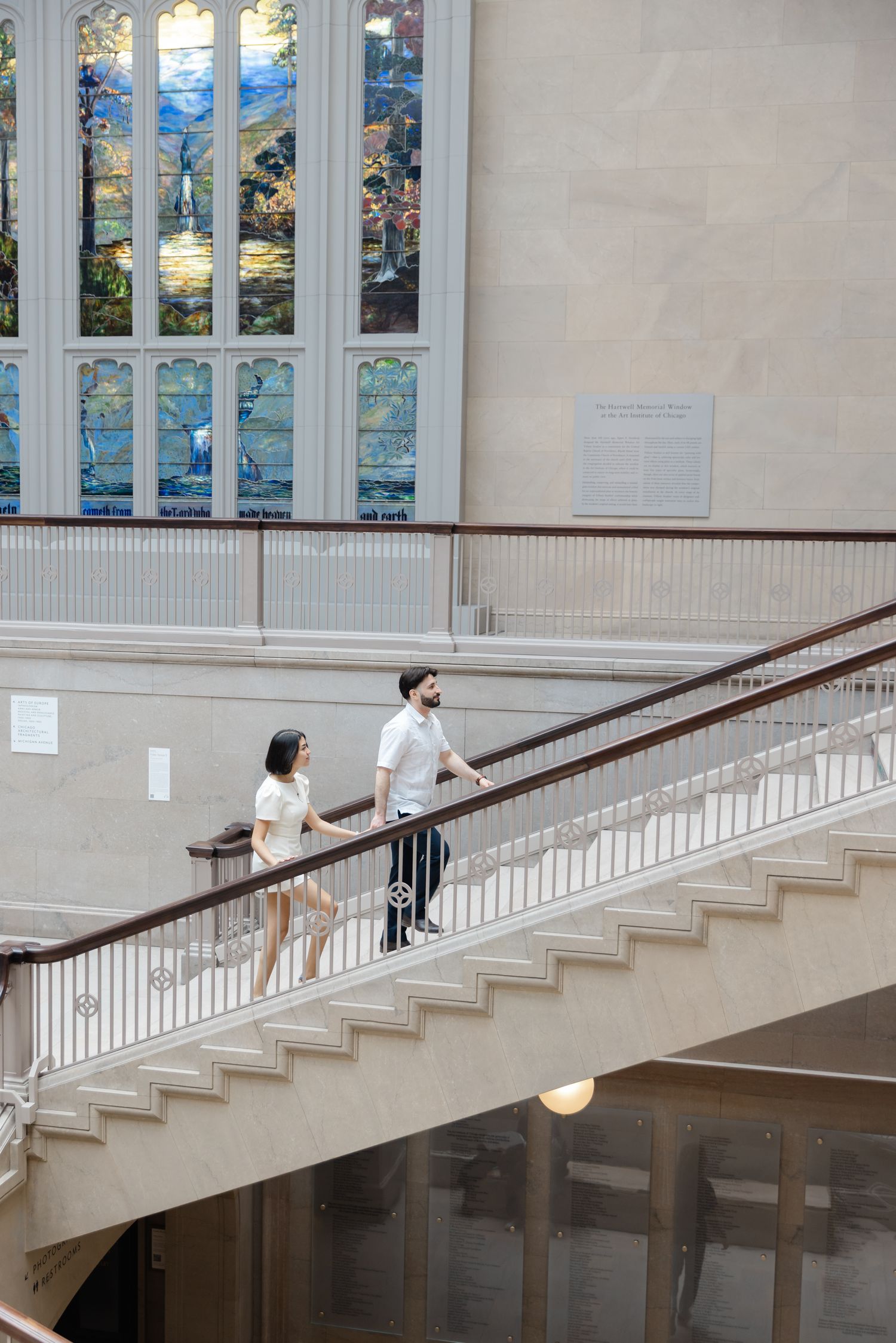 Couple walking up grand staircase with colorful stained glass window in elegant museum interior.