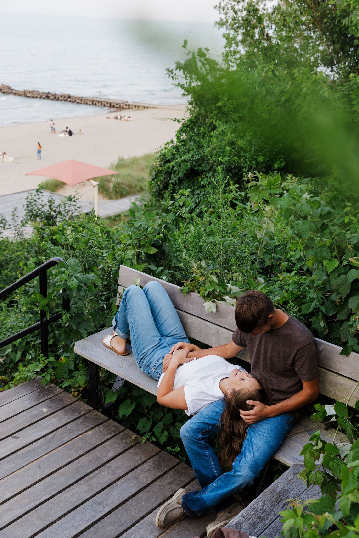 Couple relaxing on a wooden bench overlooking a beach, surrounded by lush greenery on a scenic overlook.