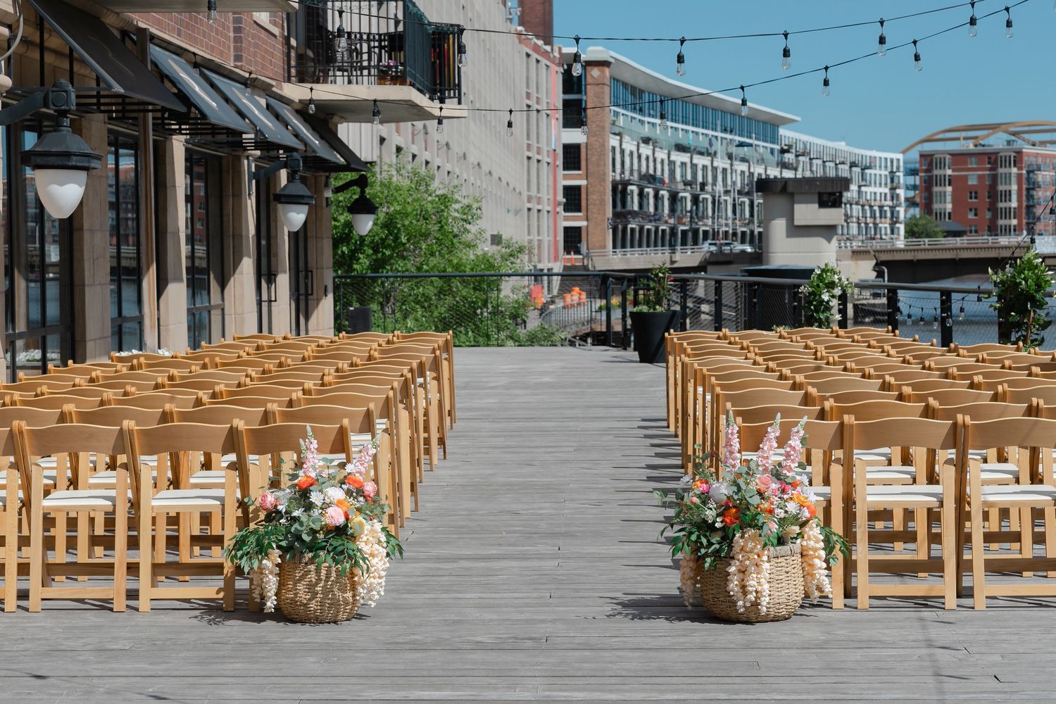 Outdoor wedding ceremony setup with wooden chairs, floral arrangements, string lights, and urban skyline backdrop.