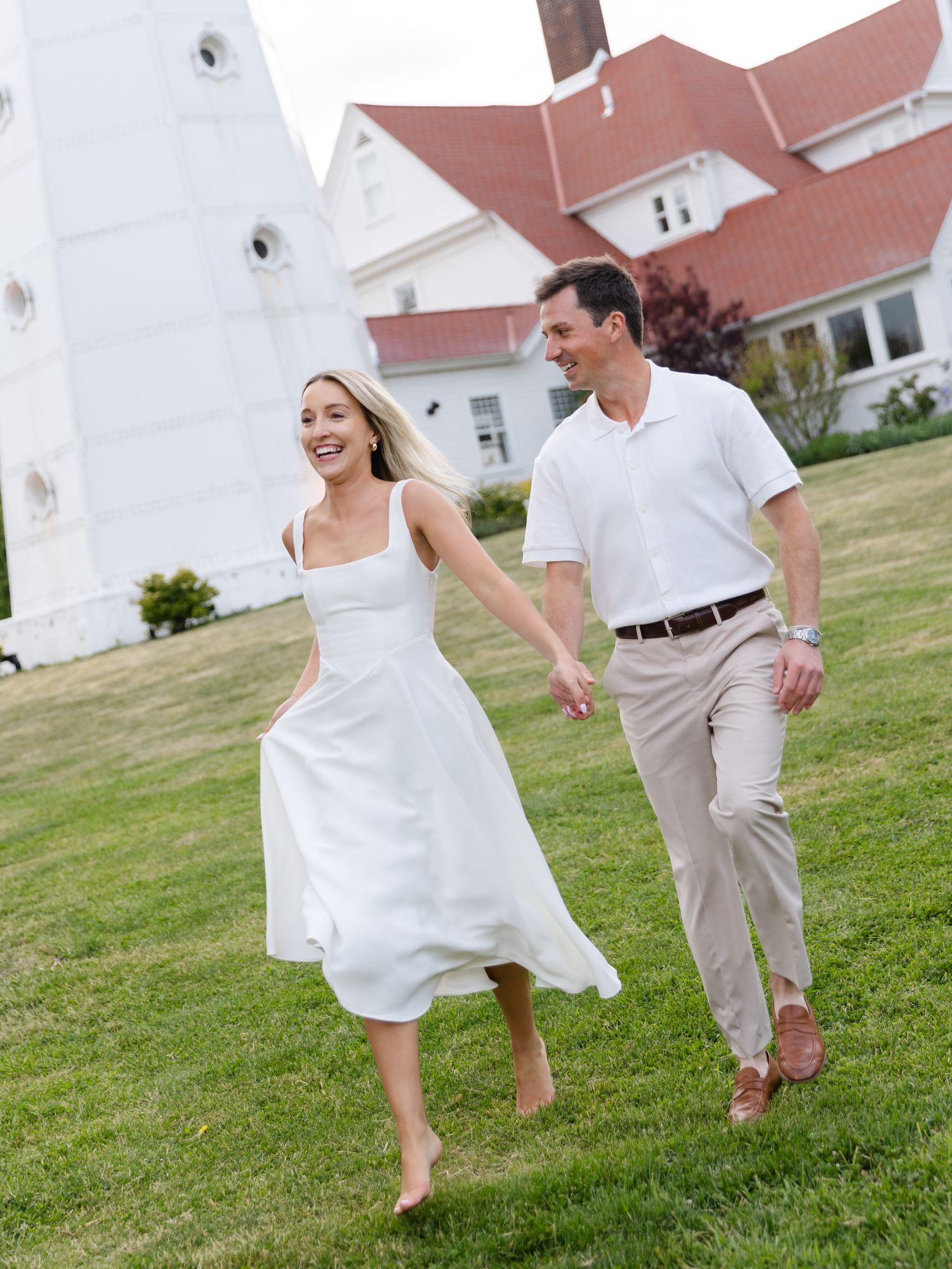 Happy couple holding hands and running on green lawn near white lighthouse and red-roofed house.