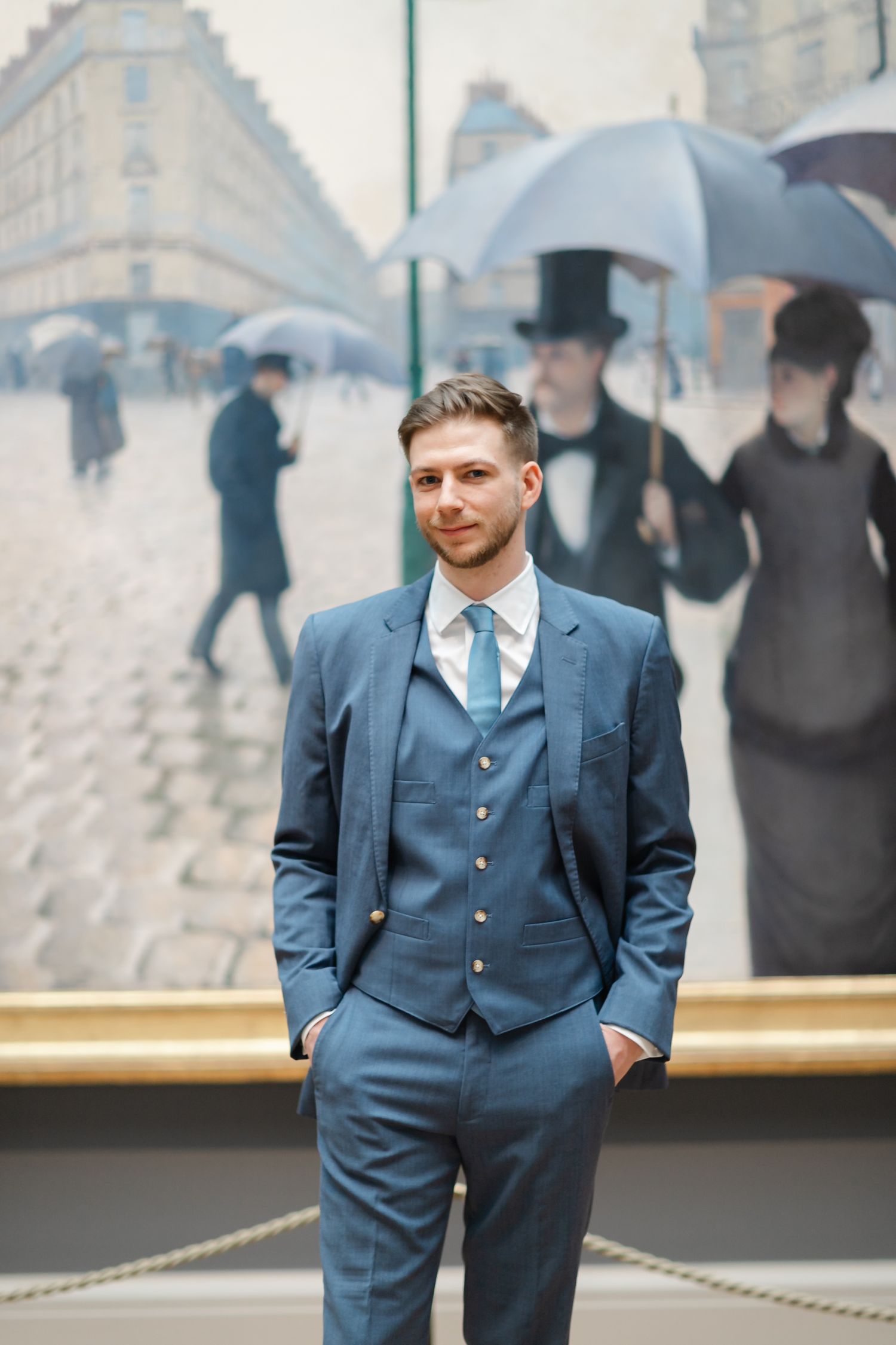 Man in blue three-piece suit standing before Caillebotte's Paris Street Rainy Day painting in museum.