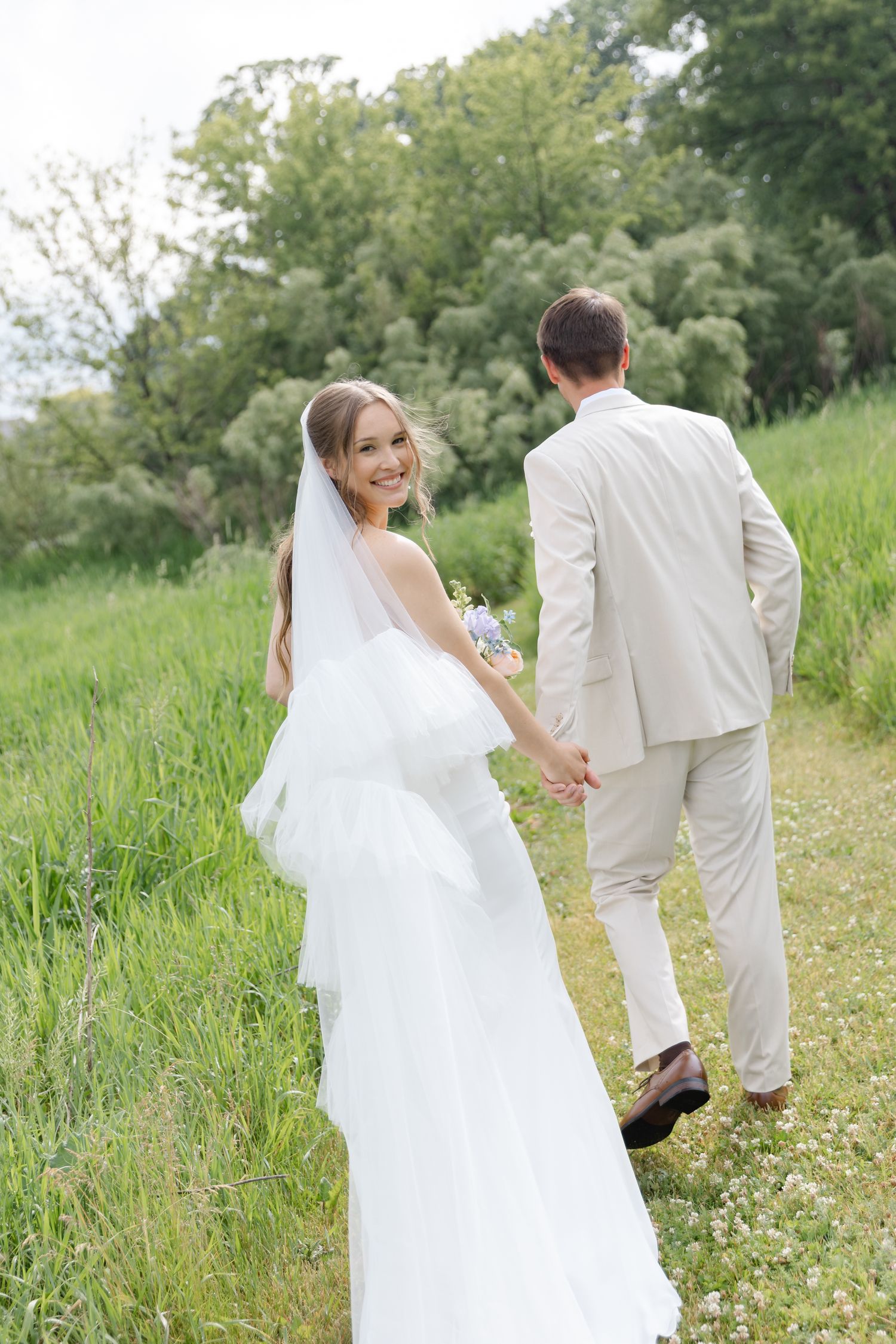 Smiling bride in white gown and veil holds groom's hand walking on green path, glancing back at camera.