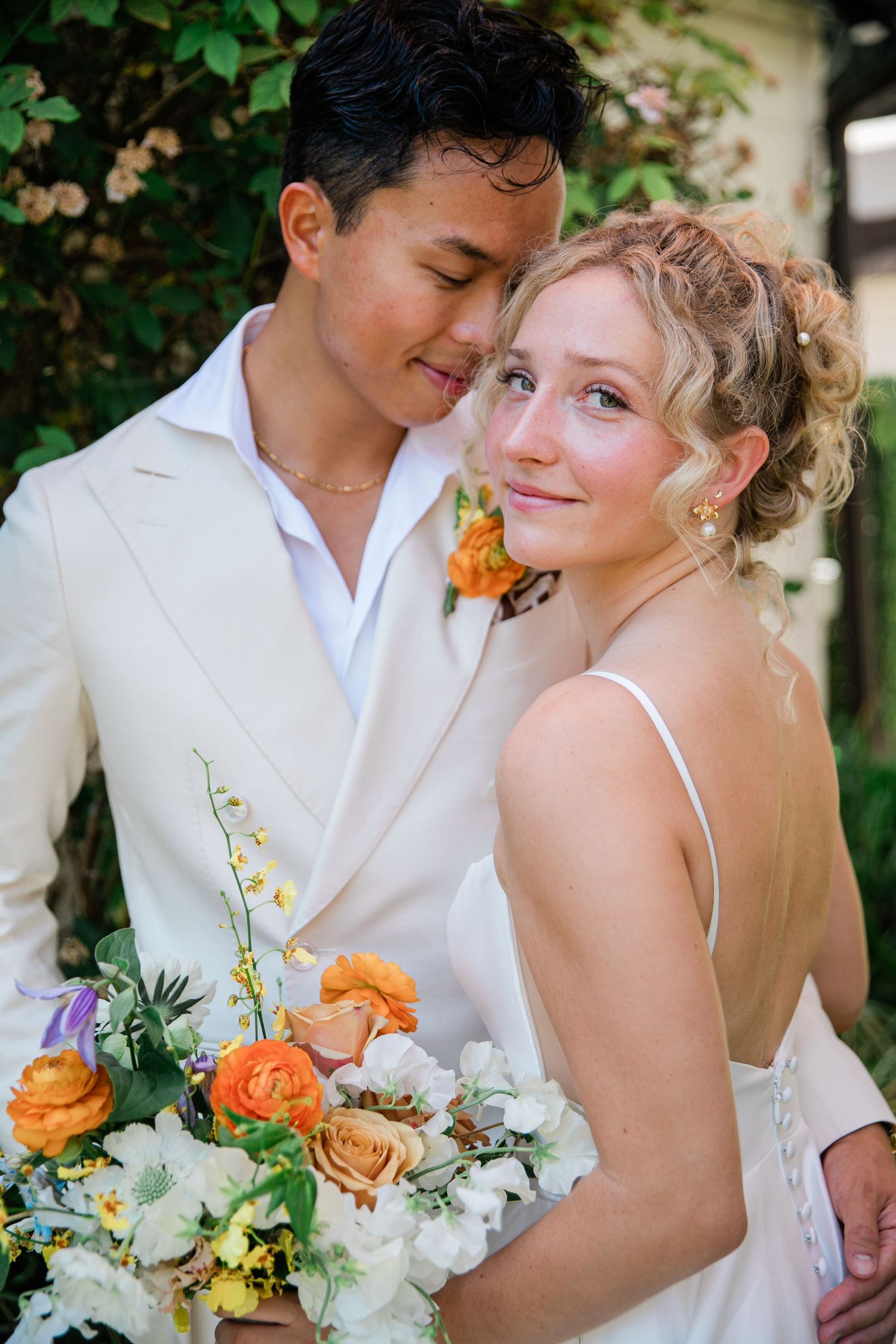 A romantic wedding couple embraces outdoors while holding a vibrant orange and white floral bouquet.