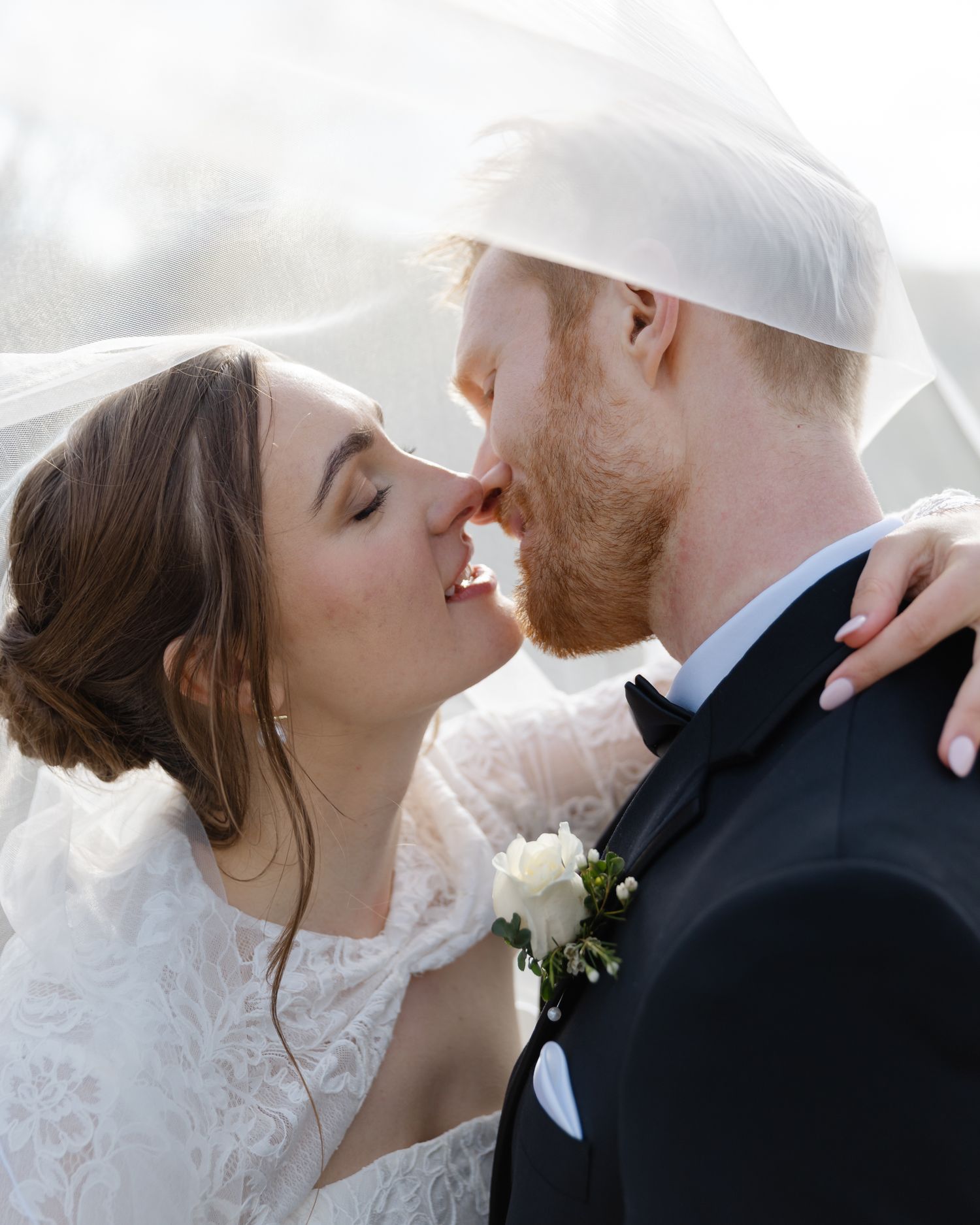 Bride and groom sharing a romantic kiss under a flowing white veil on their wedding day.
