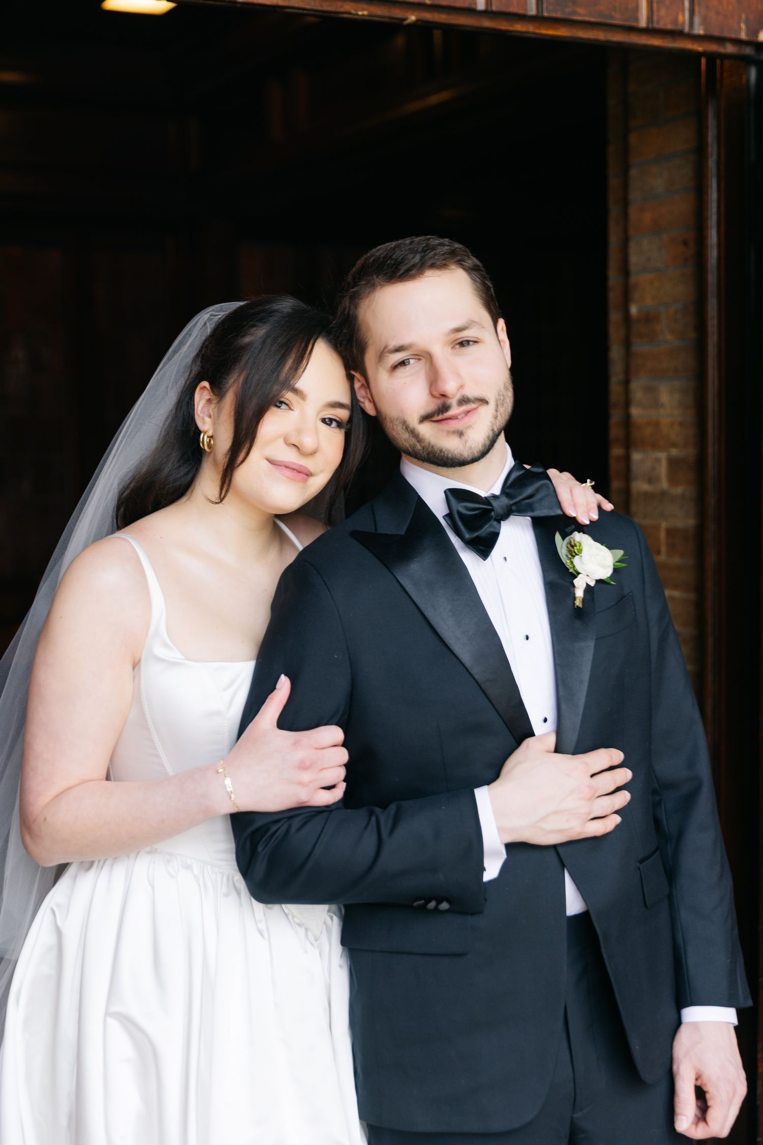 Elegant bride in white gown and veil embraces groom in black tuxedo with bow tie and boutonniere.