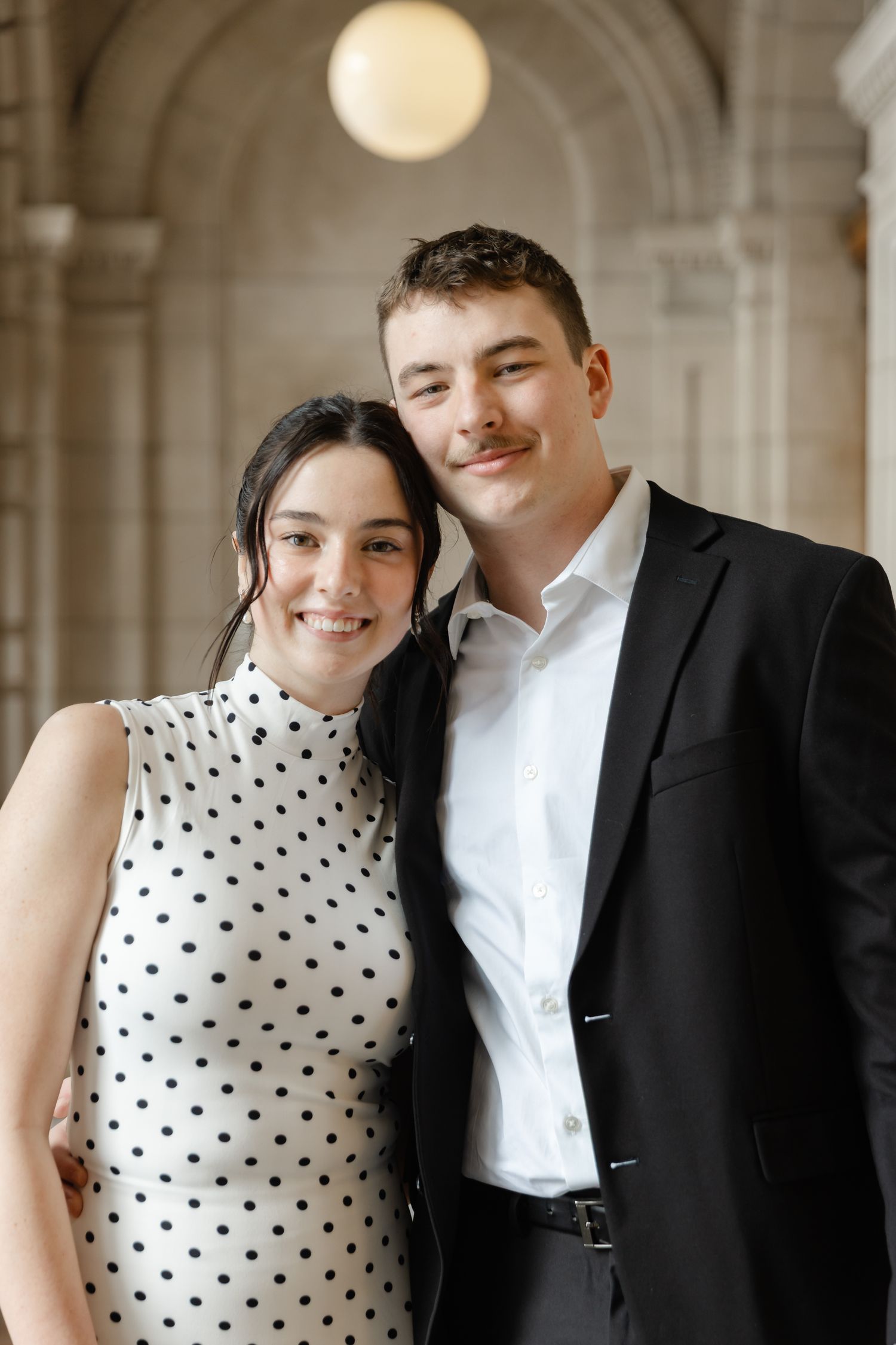 Smiling couple in formal attire, woman in polka dot dress and man in black suit, posing together in elegant venue.