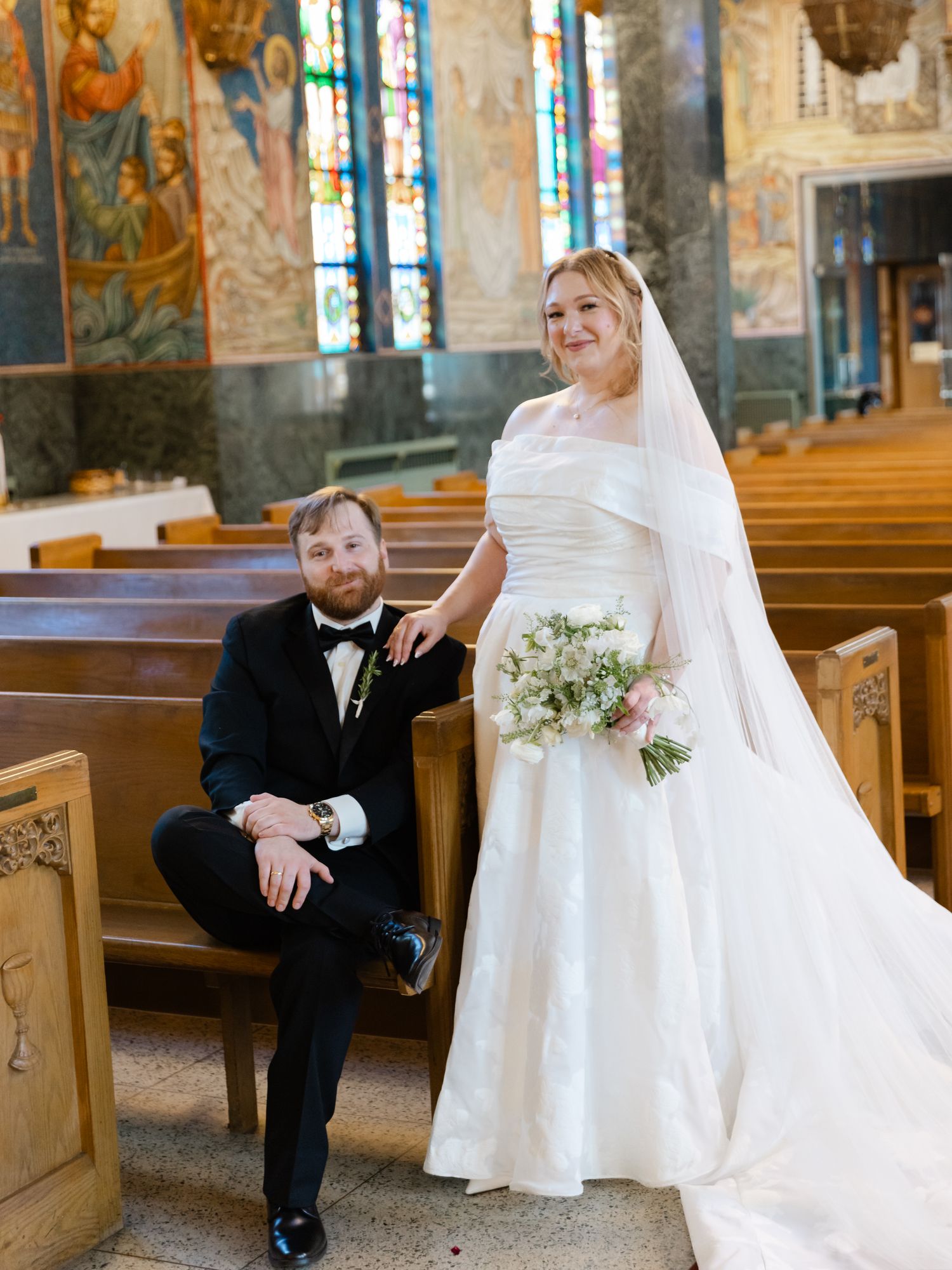 Bride in white gown with veil and groom in black tuxedo pose inside a church with stained glass windows.