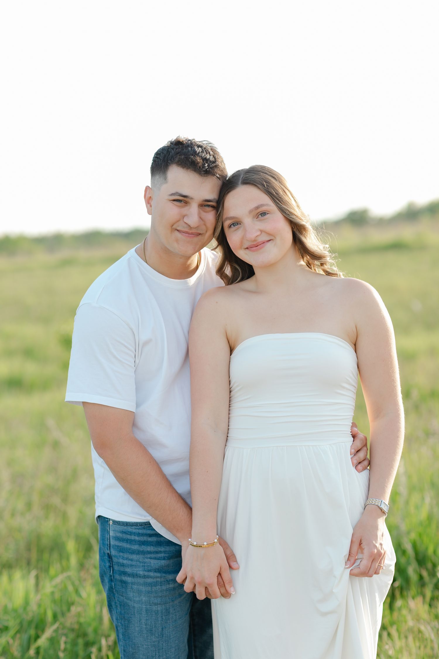 Happy couple posing in green field, man in white tee and jeans, woman in white strapless dress.