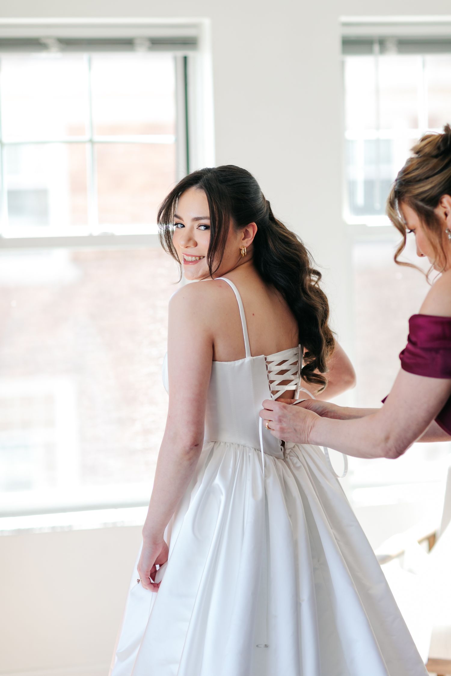 Bride in white lace-up gown smiles over shoulder as bridesmaid in burgundy dress fastens her wedding dress.