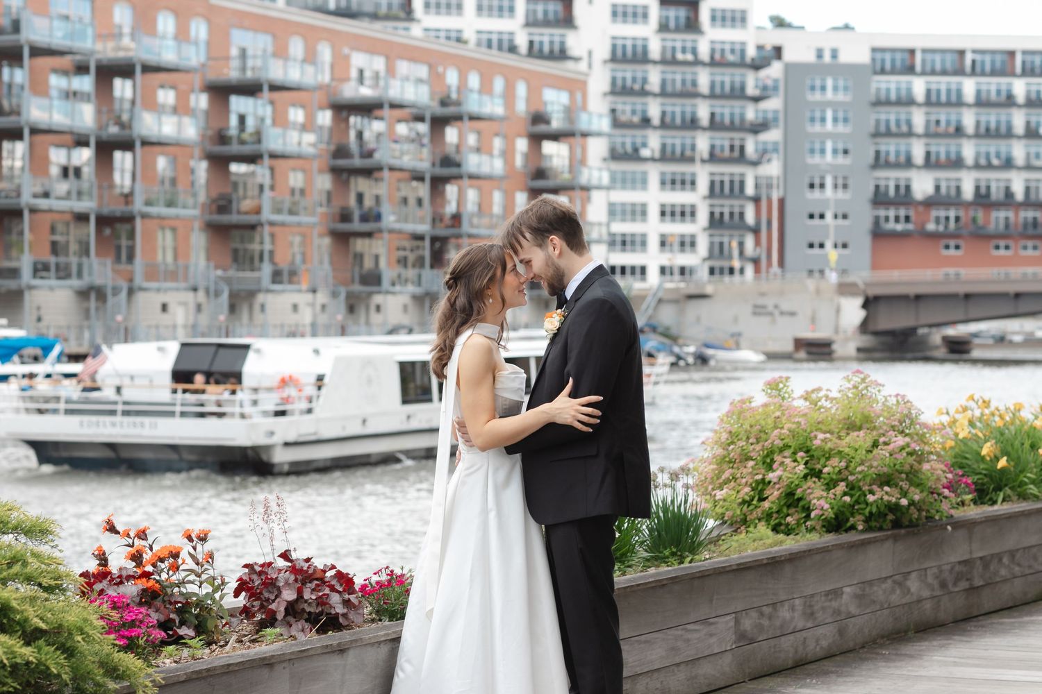 Bride and groom share a romantic kiss by a waterfront with boats and city buildings in the background.