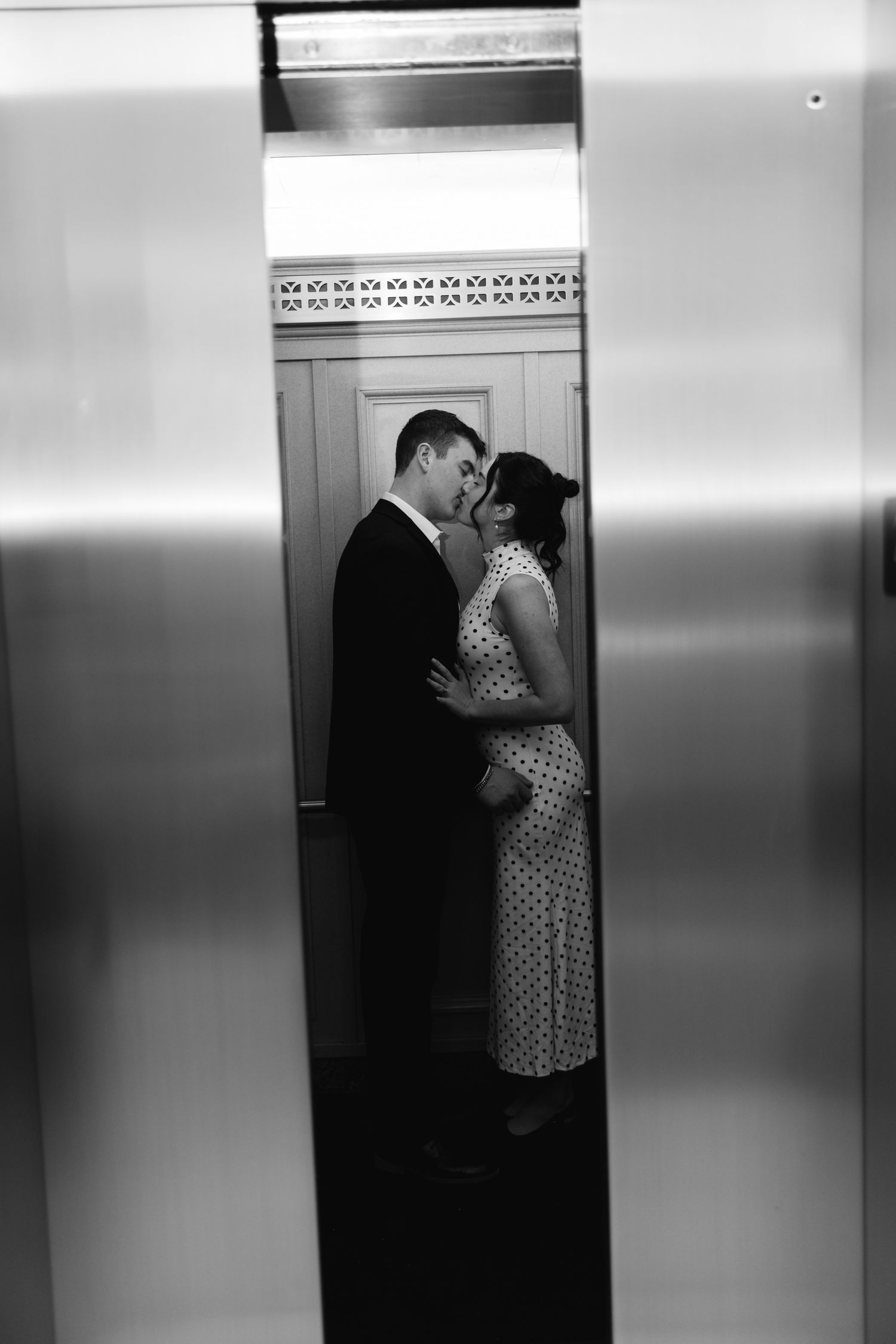 Romantic couple sharing an intimate moment in a sleek elevator, woman in polka dot dress, black and white photo.