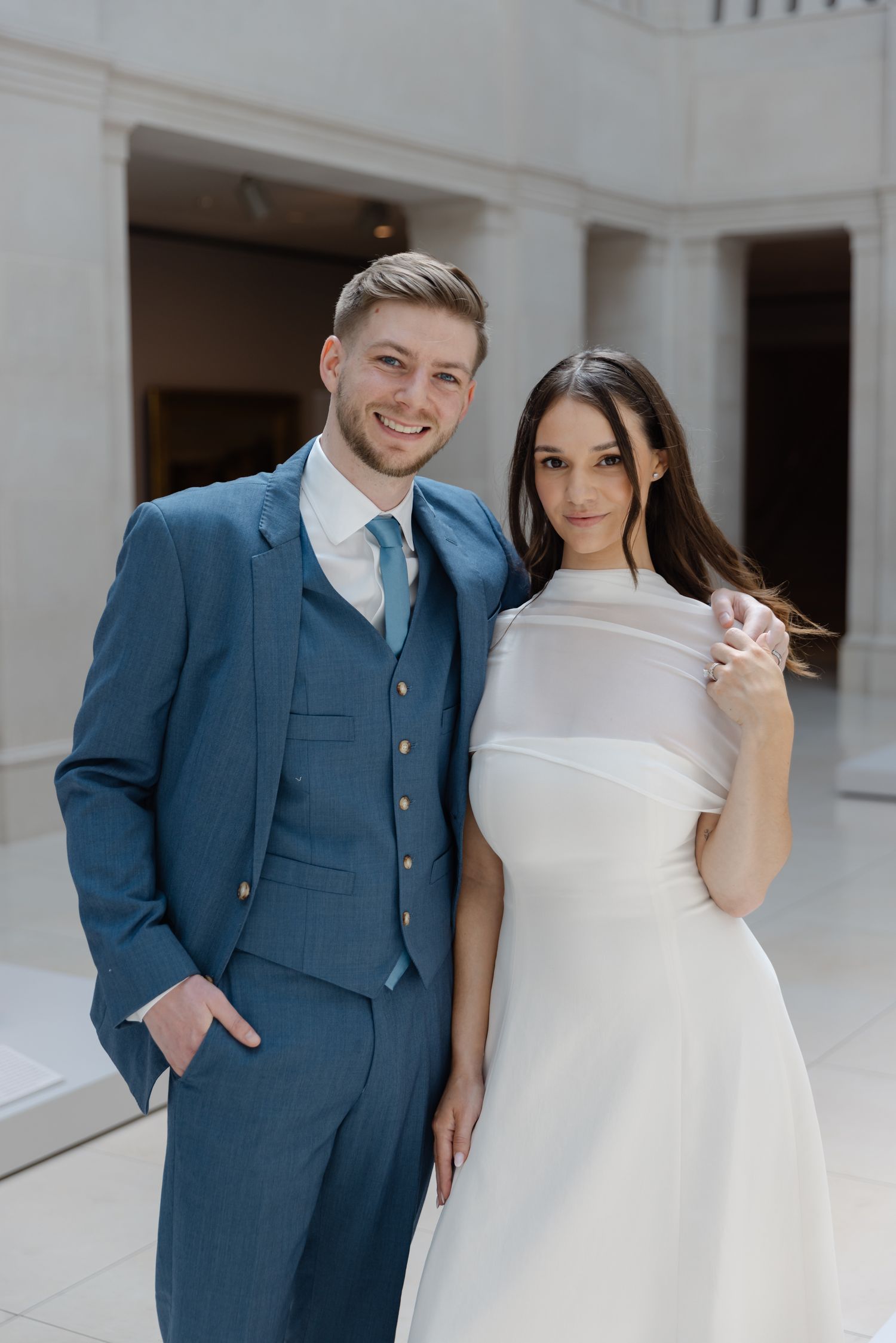 Smiling couple in wedding attire, man in blue suit and woman in white dress, posing in elegant venue.