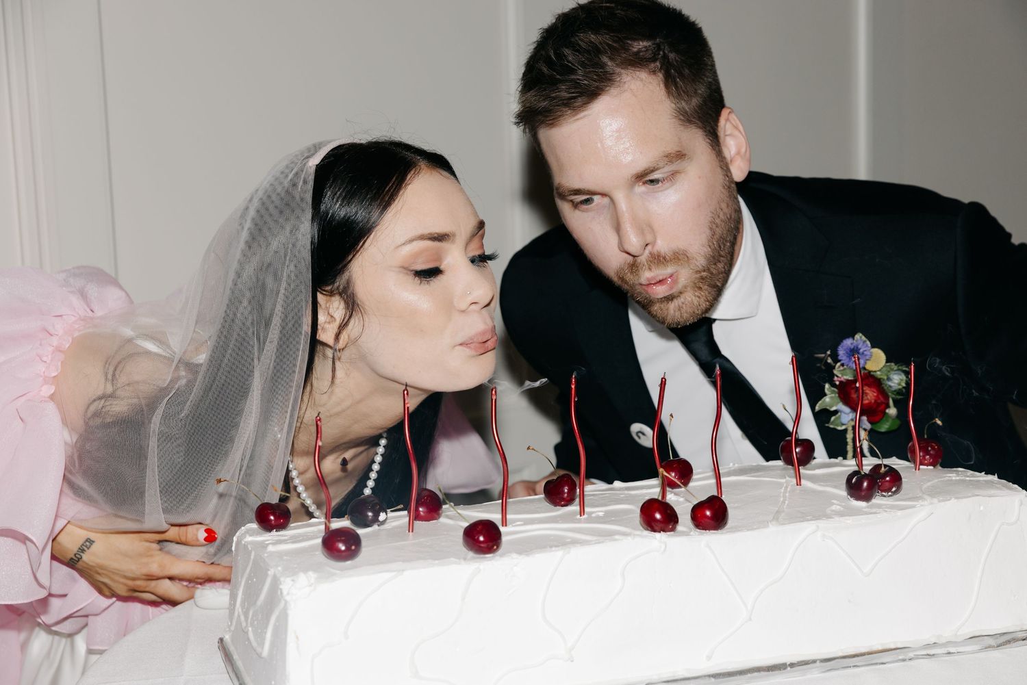 Bride and groom blowing out candles on white wedding cake decorated with fresh cherries.