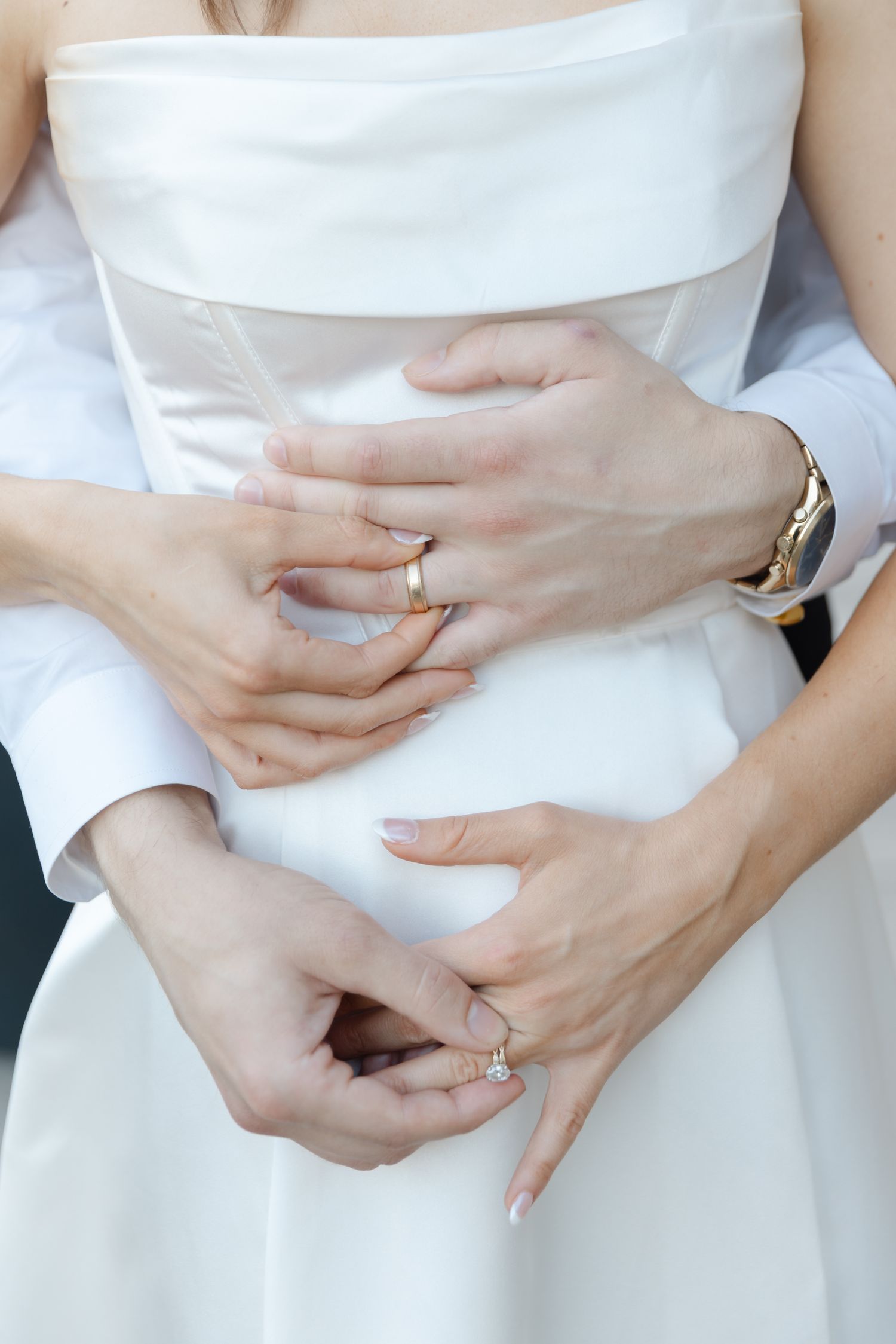 Close-up of newlyweds hands with gold wedding rings and diamond engagement ring on brides white gown.