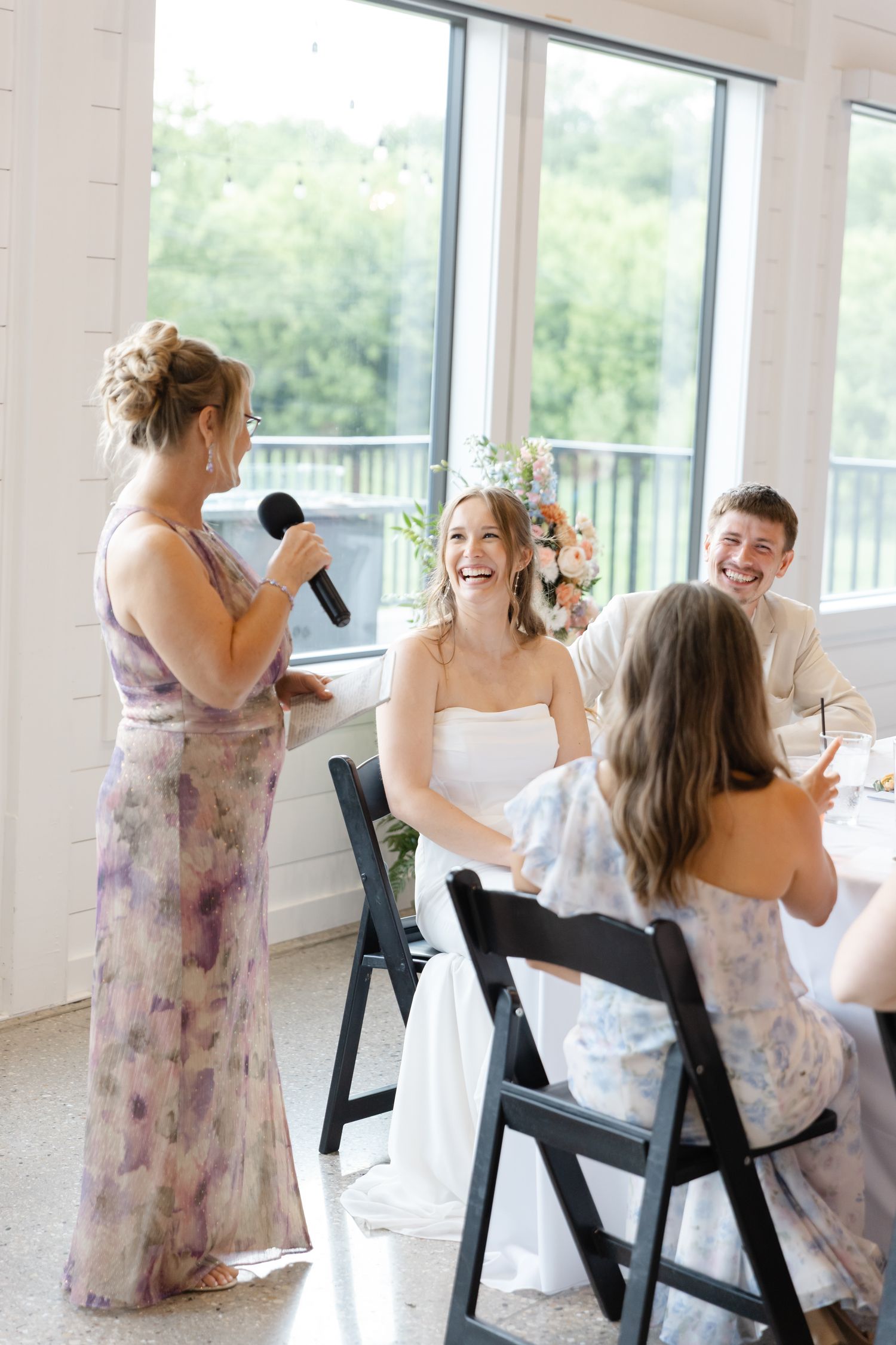 Bridesmaid gives a heartfelt speech with microphone as bride laughs joyfully at elegant wedding reception.