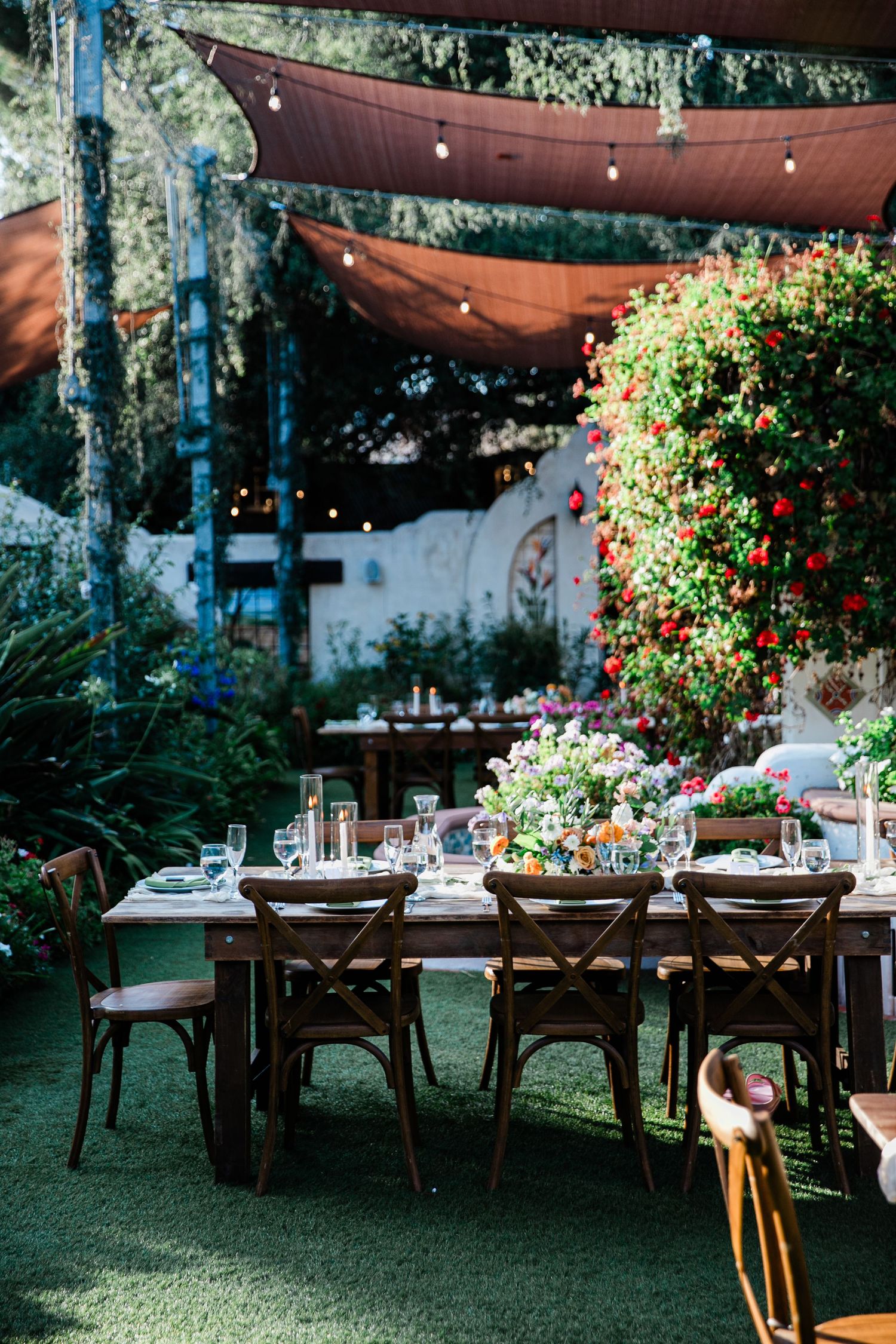 An elegant outdoor wedding reception setup with wooden tables under brown fabric canopies and string lights.
