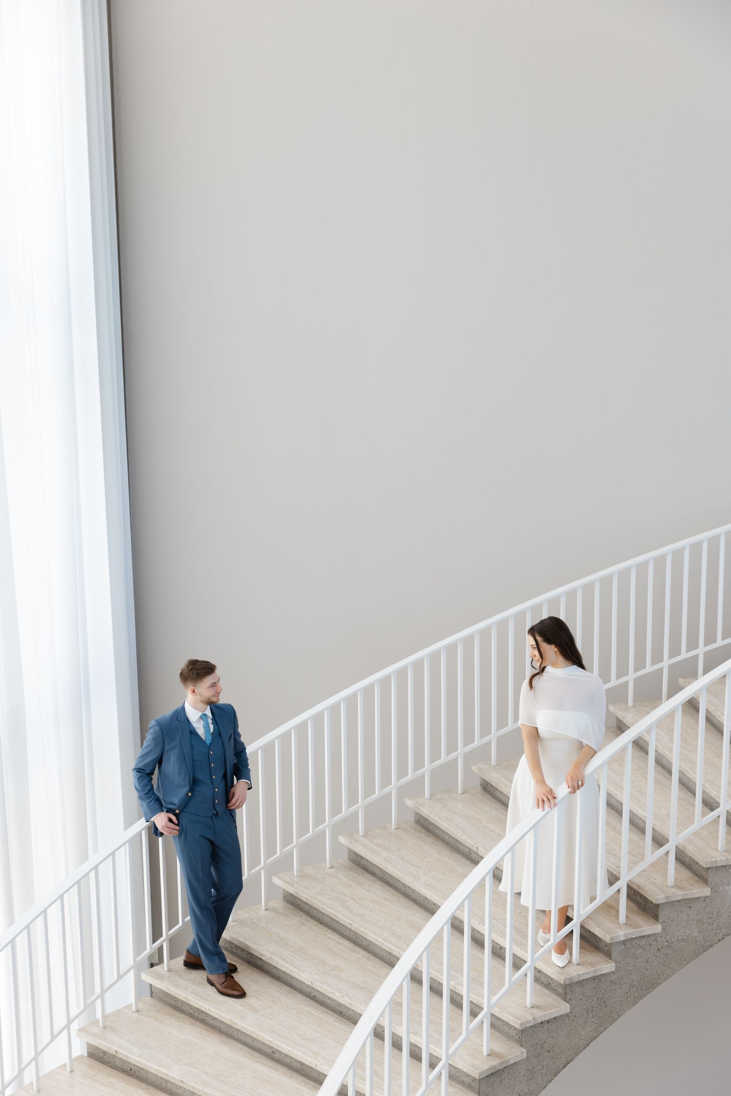 Bride and groom on elegant curved white staircase, man in blue suit, woman in white dress, modern interior.