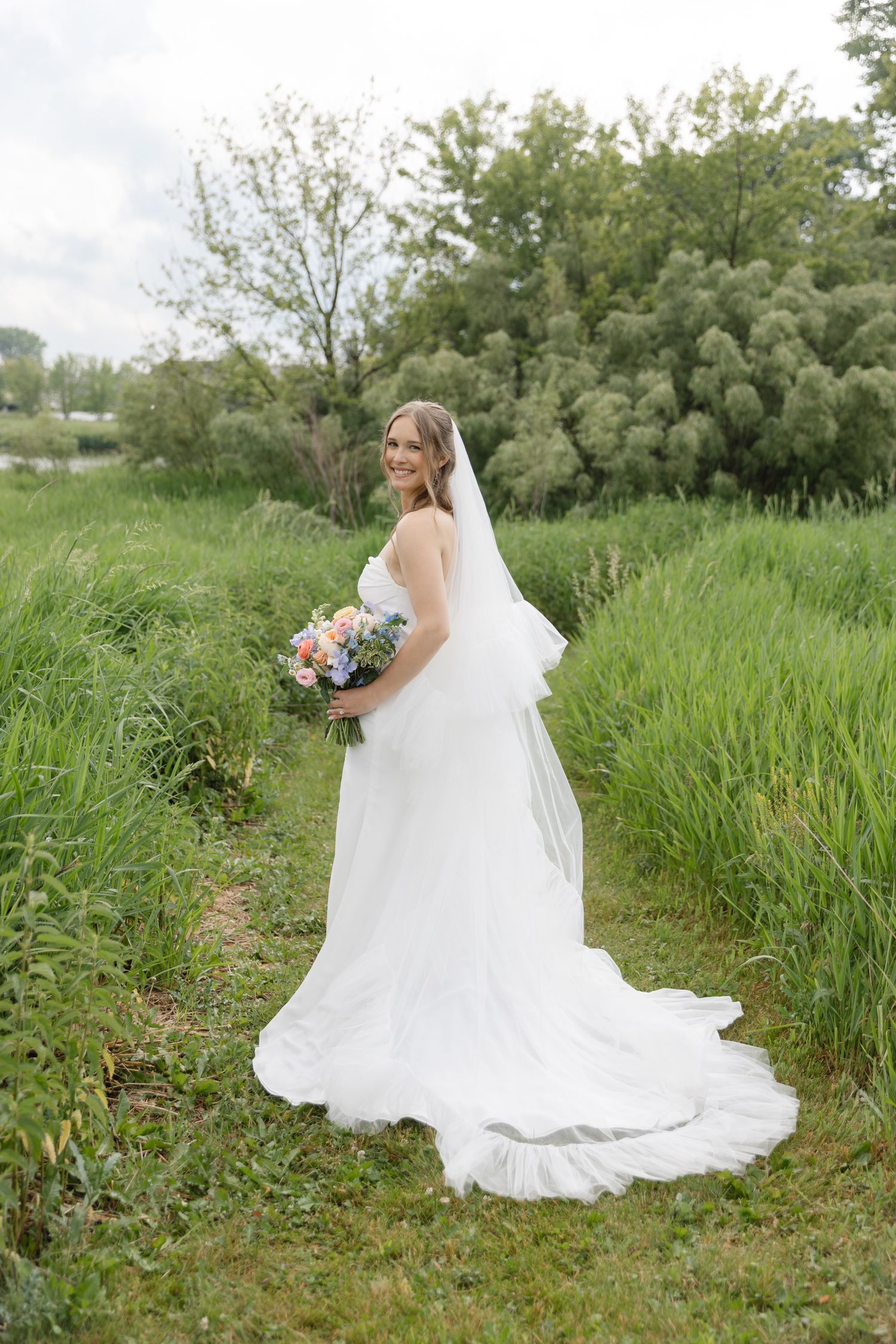 Smiling bride in white gown and veil holds colorful bouquet while standing on a lush green garden path.