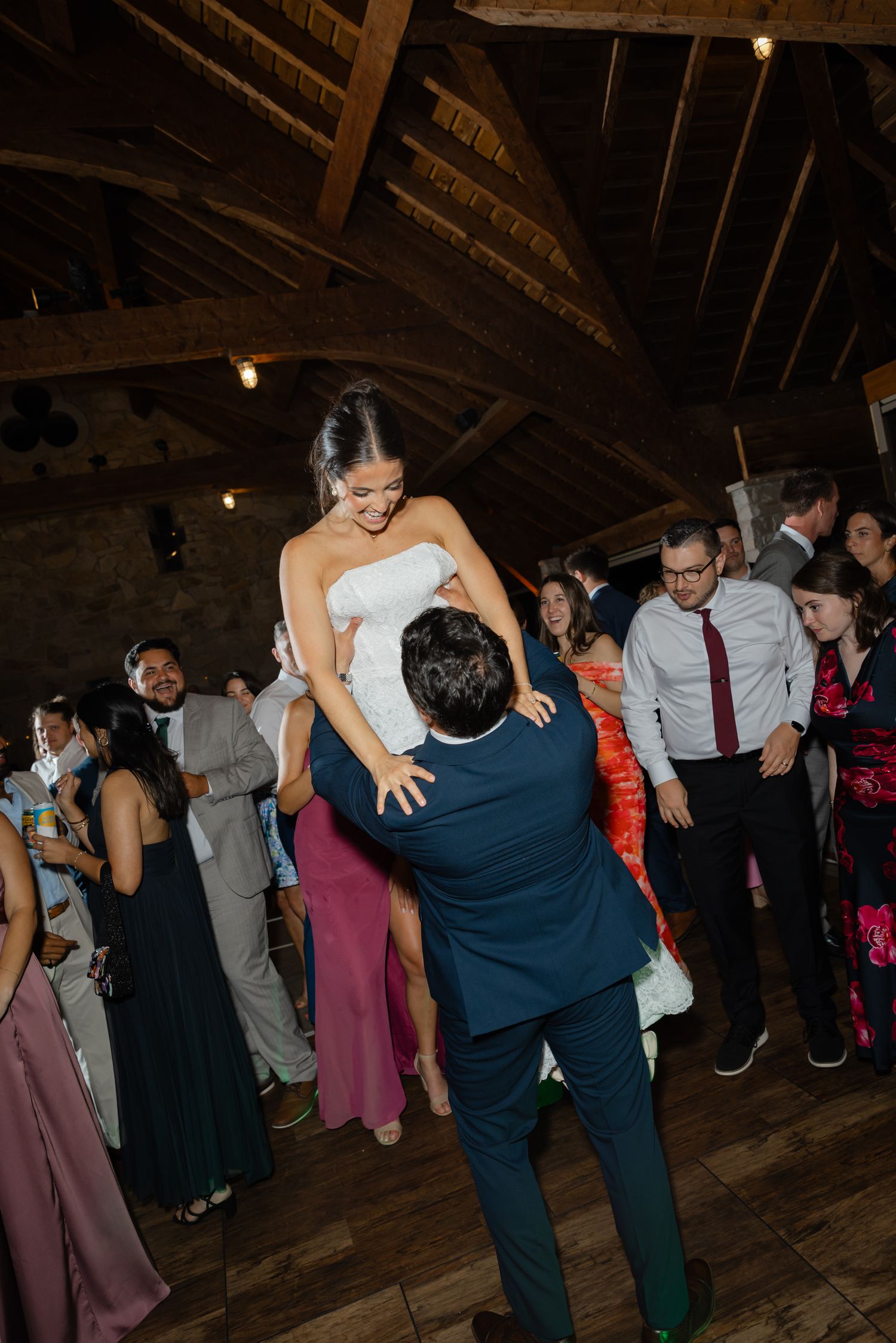 Groom lifting bride in white dress on wedding reception dance floor in rustic barn venue with guests watching.